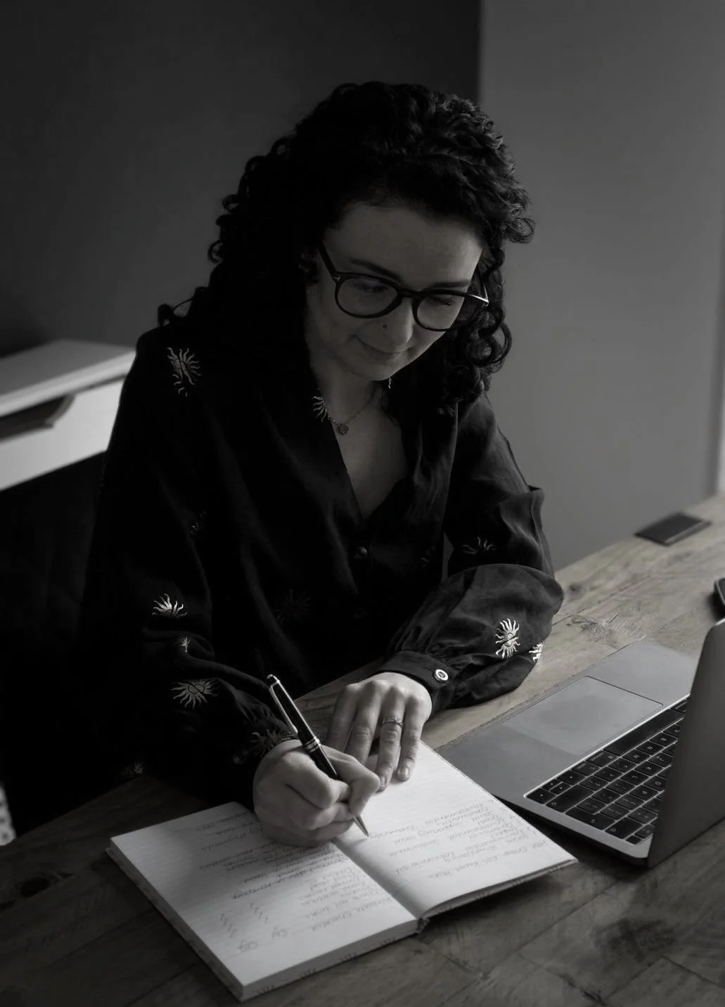 Woman with dark curly hair and glasses writing in a notebook with a laptop on a wooden desk