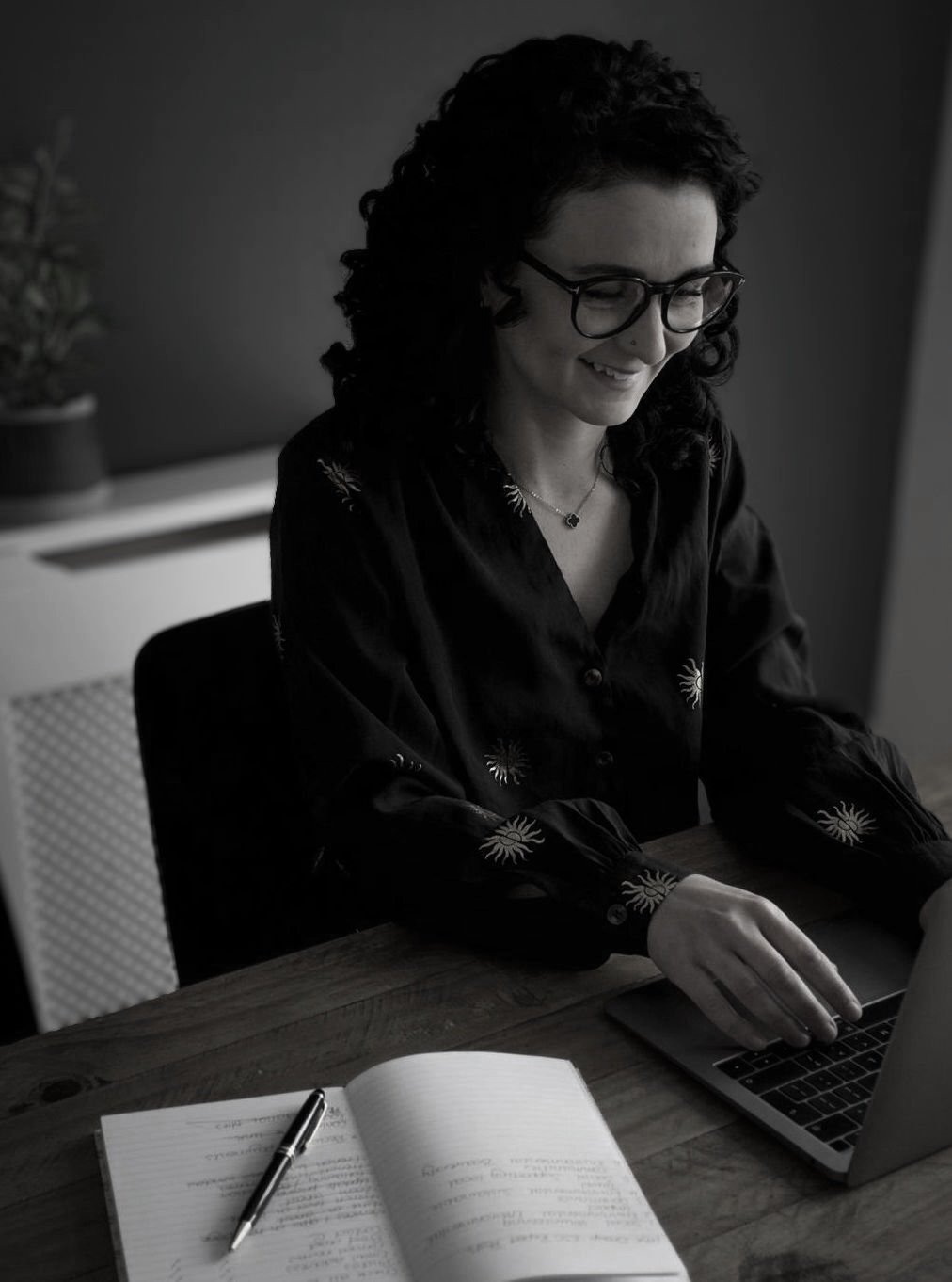 Woman with curly hair and glasses typing on a laptop at a wooden desk with an open notebook and pen.