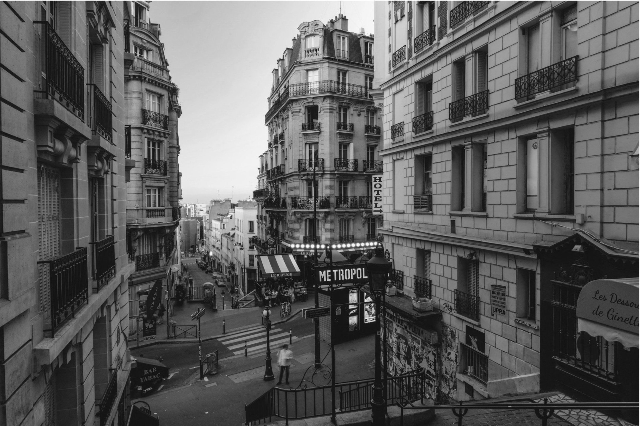 Black and white photo of a French street view with vintage buildings, ornate balconies, a "METROPOL" sign, hotel signage, and cobbled sidewalks.