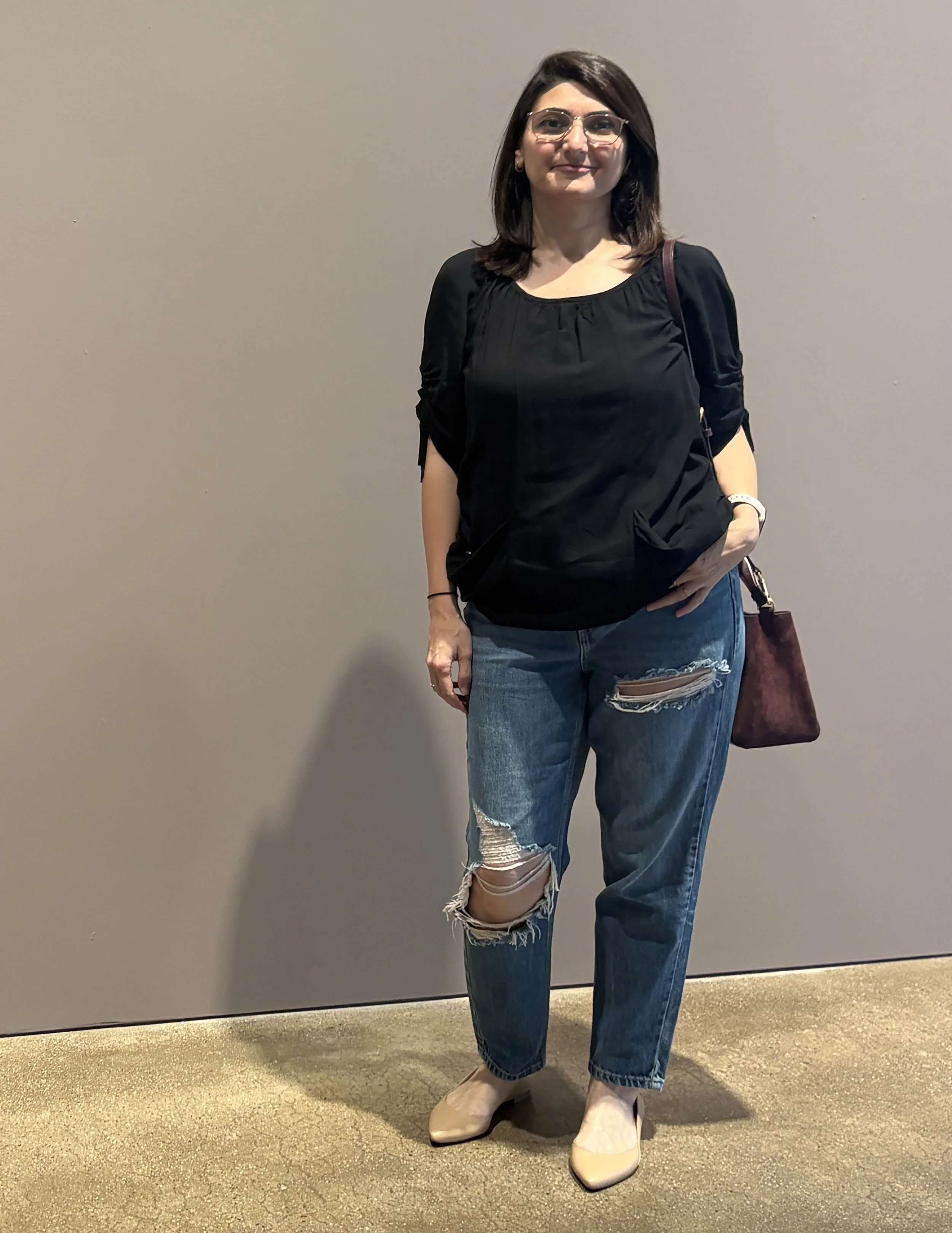 A woman standing in front of a plain wall, wearing glasses, a black blouse, distressed jeans, and beige shoes, holding a brown handbag.