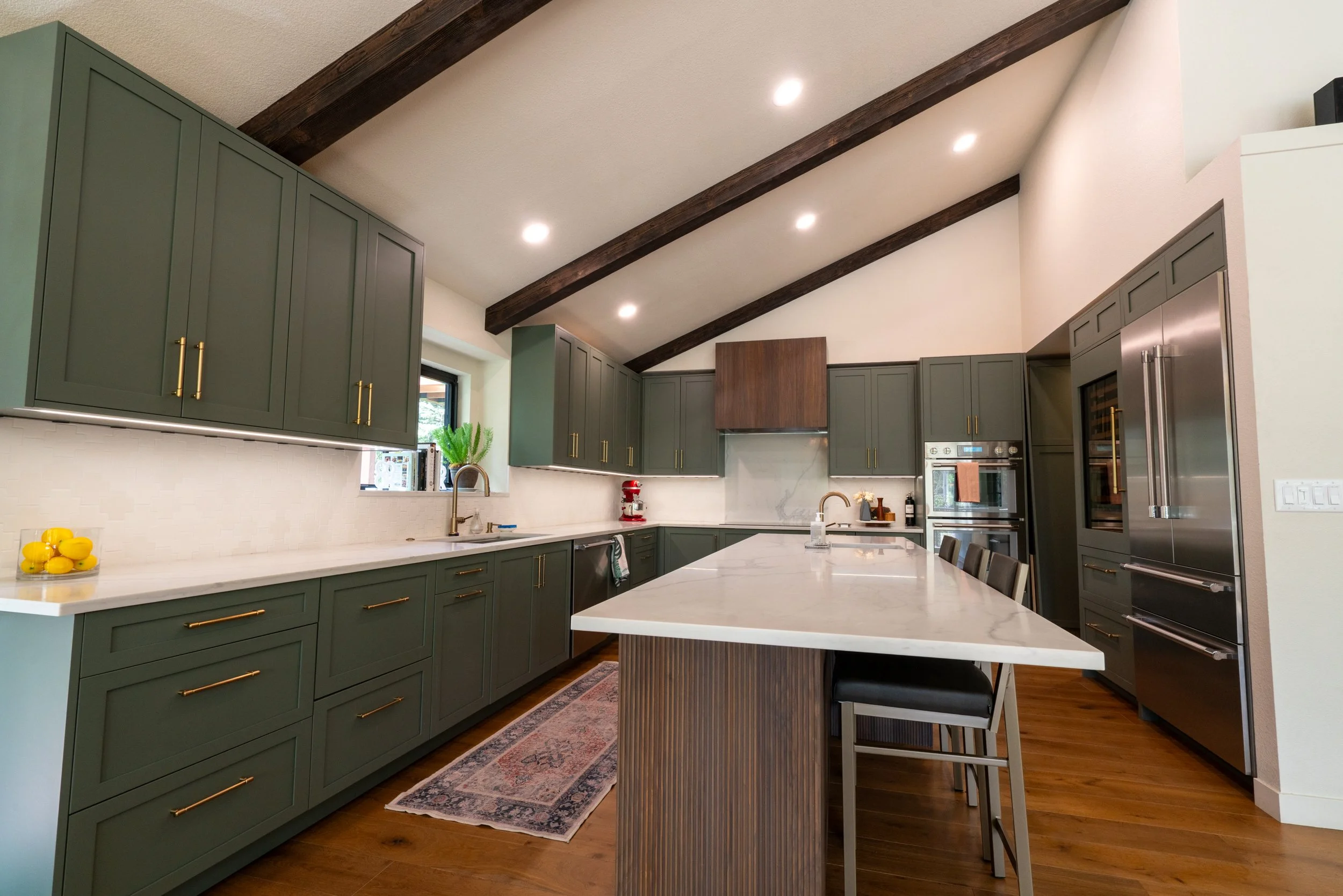 Modern kitchen with green cabinets, white marble countertops, stainless steel appliances, and a large island with seating. Dark wood beams run across the ceiling, and there is recessed lighting. A window allows natural light into the space.