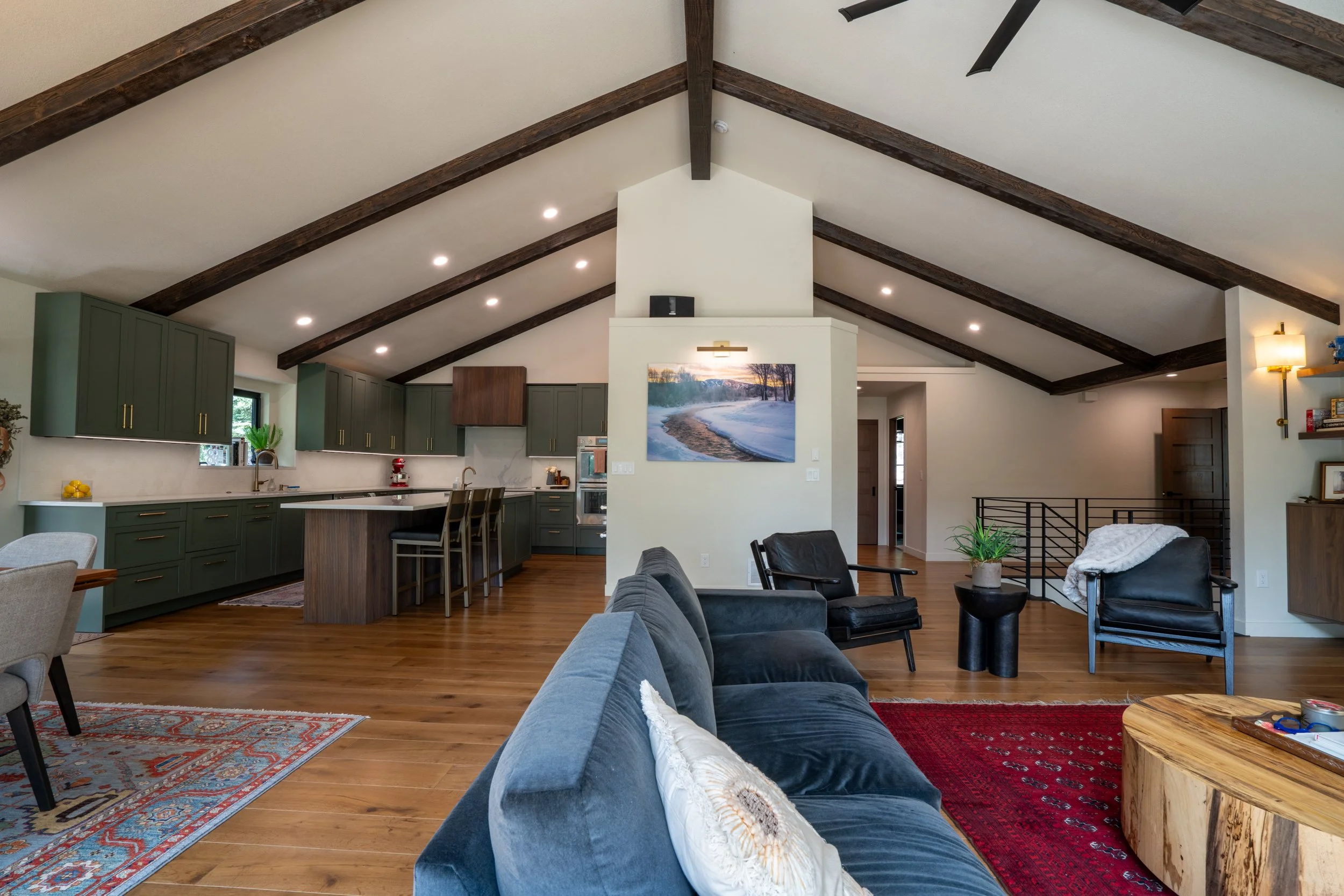 Open-concept living room and kitchen with vaulted ceiling, exposed dark wood beams, green cabinets, and hardwood floors. Furniture includes a blue velvet sofa, black armchairs, and a round wooden coffee table. There is a wall-mounted painting of a winter landscape, with recessed ceiling lights and indoor plants.