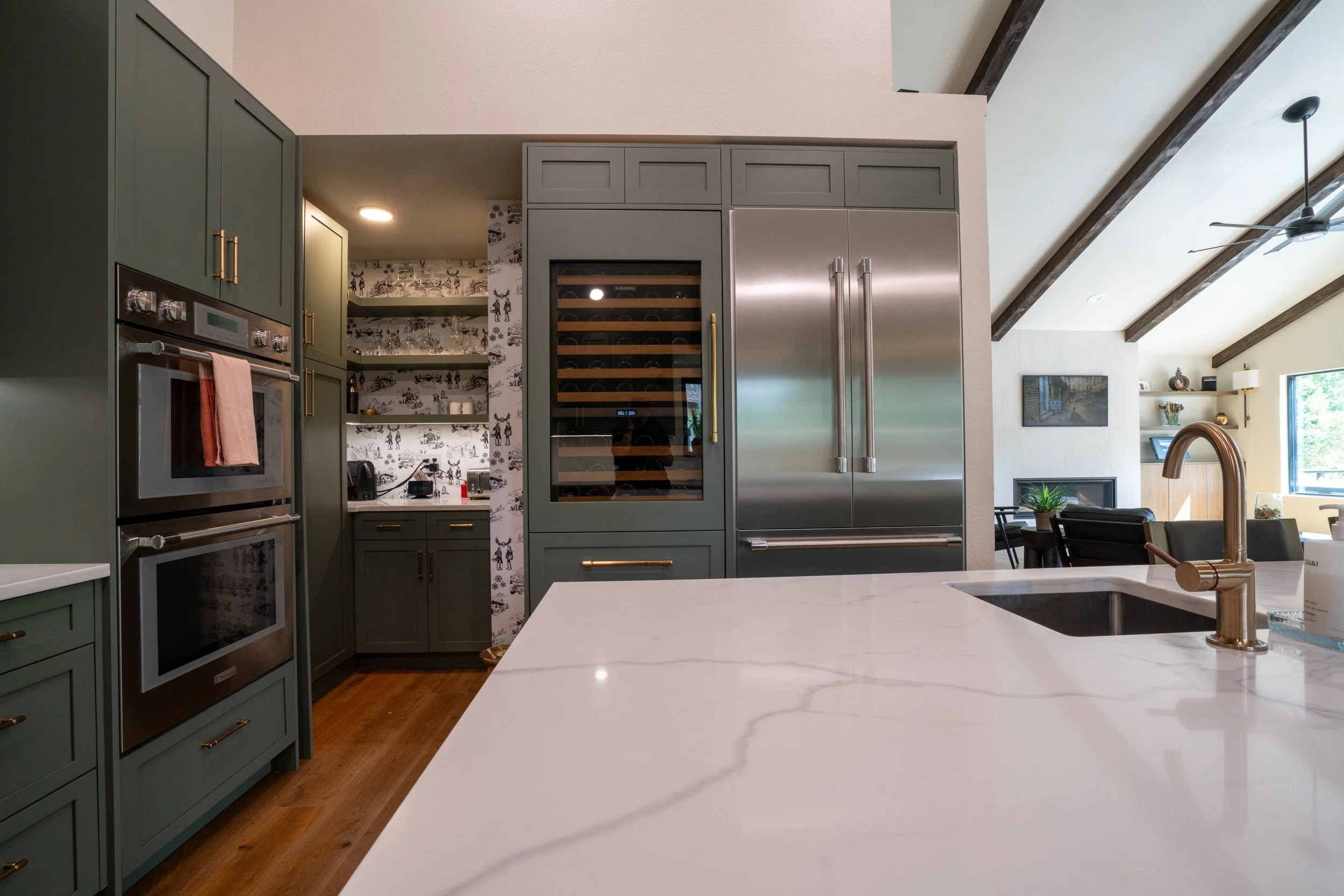Modern kitchen with green cabinets, stainless steel appliances, a wine cooler, and a white marble countertop island, overlooking a living area with a TV and large windows.
