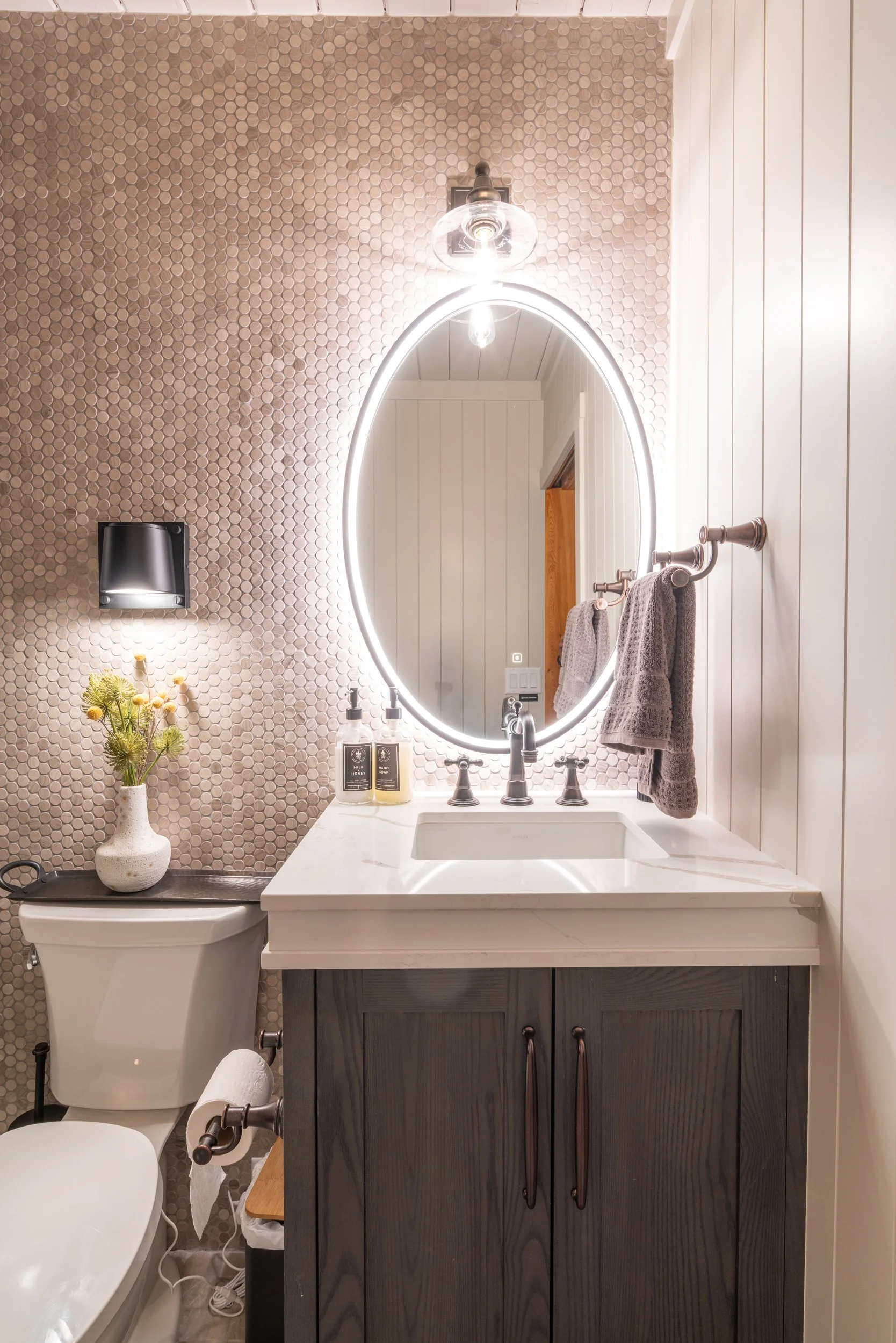 Bathroom with a vanity mirror, sink, dark wood cabinet, towel hooks with towels, decorative plant, soap dispensers, light beige hexagonal tile wall, toilet, and wall-mounted TV.