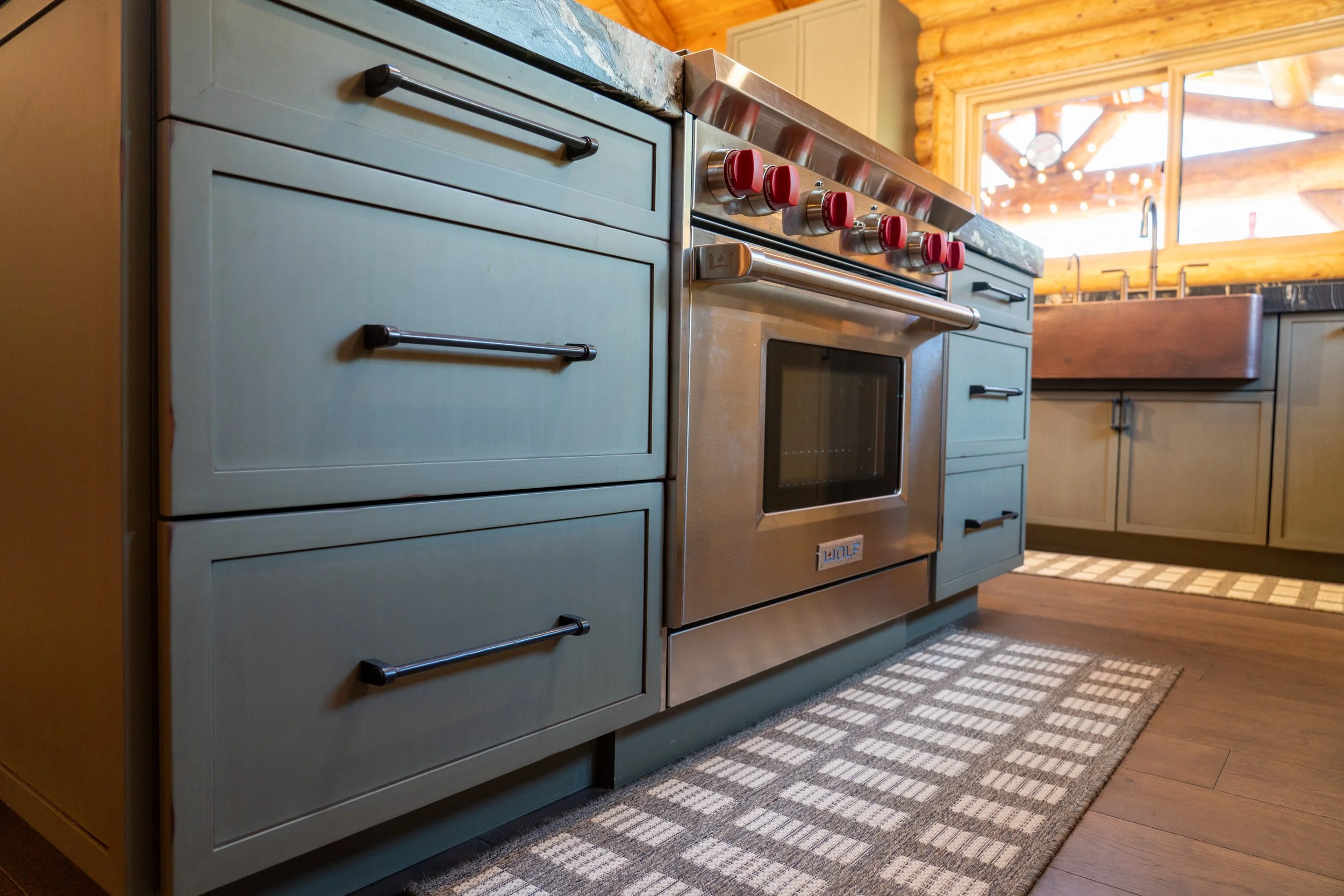 A kitchen with a stove featuring five red control knobs, a stainless steel oven, light blue cabinets, a wooden ceiling with visible logs, and a window showing part of a ceiling fan outside.