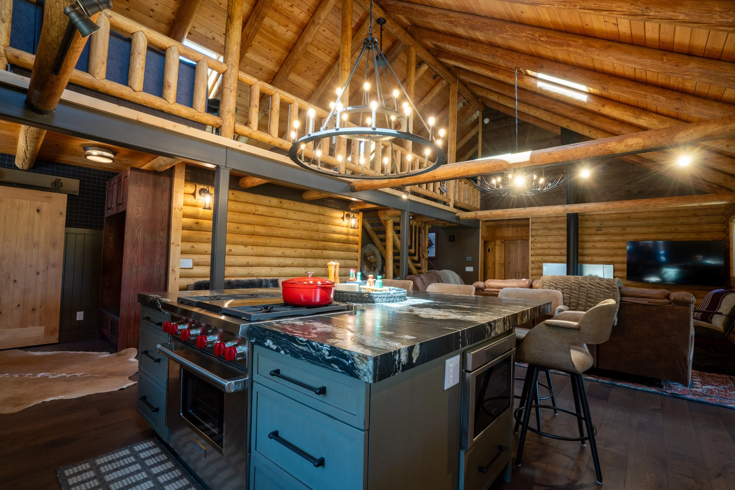 Interior of a rustic log cabin with a large kitchen island featuring a dark marble countertop, red pot, and nearby stools, living area with sofas, and wooden decor including log beams and railings.