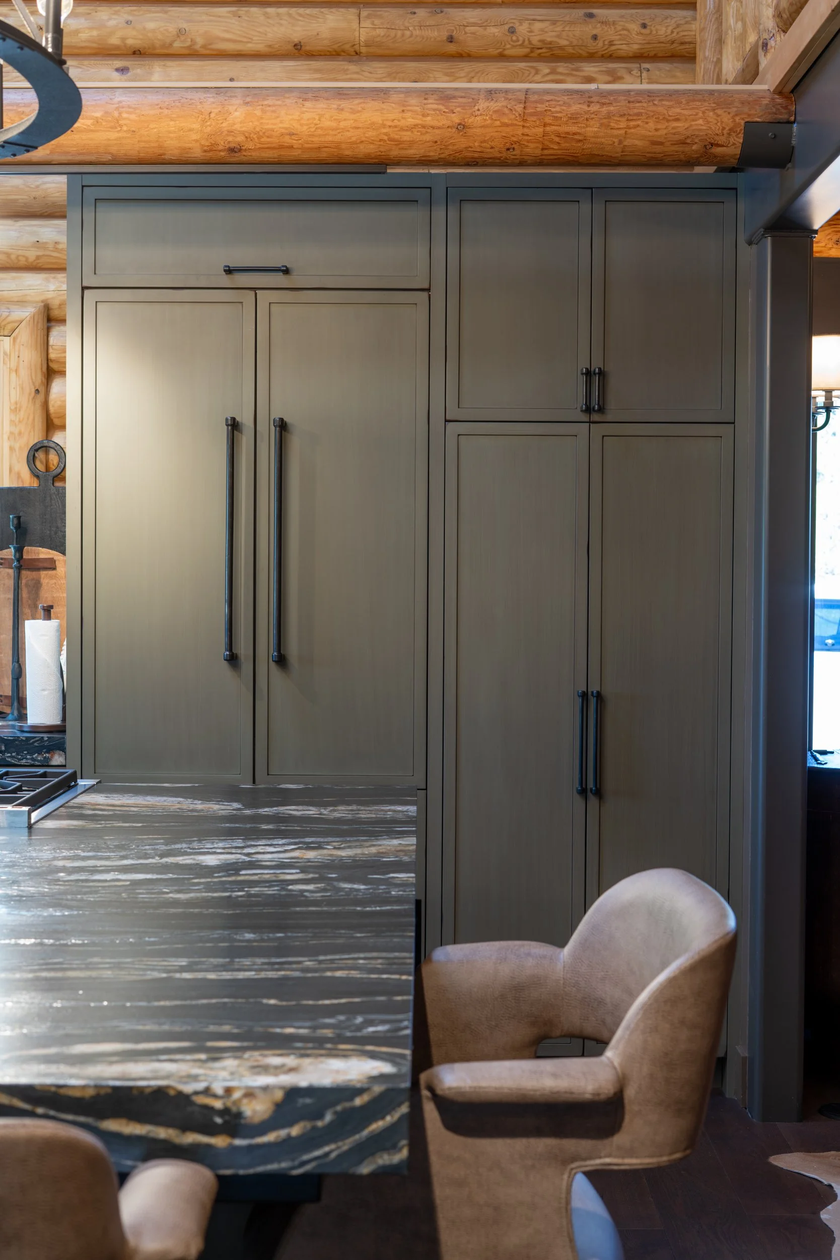 A modern kitchen with gray cabinets, wooden log walls, a marble countertop, and a beige upholstered chair.
