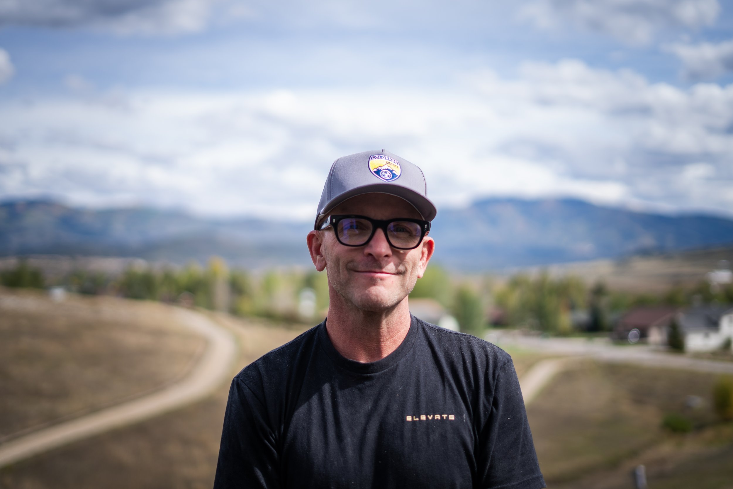 A man with glasses and a gray cap smiling outdoors under a cloudy sky.