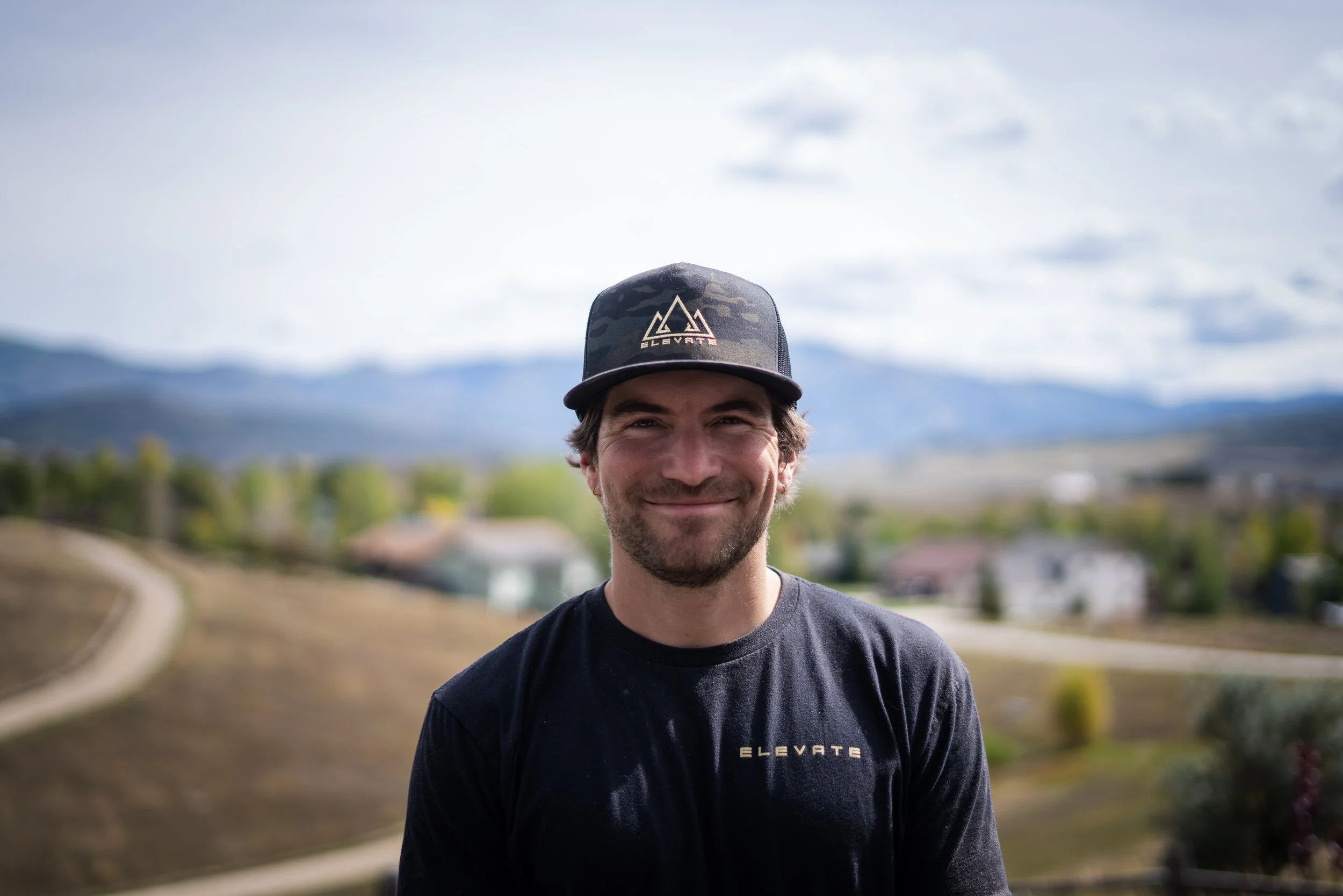 A smiling man wears a black baseball cap with a mountain logo and the word 'ELEVATE' and a black T-shirt with a similar logo, standing outdoors with a background of mountains, cloudy sky, and greenery.