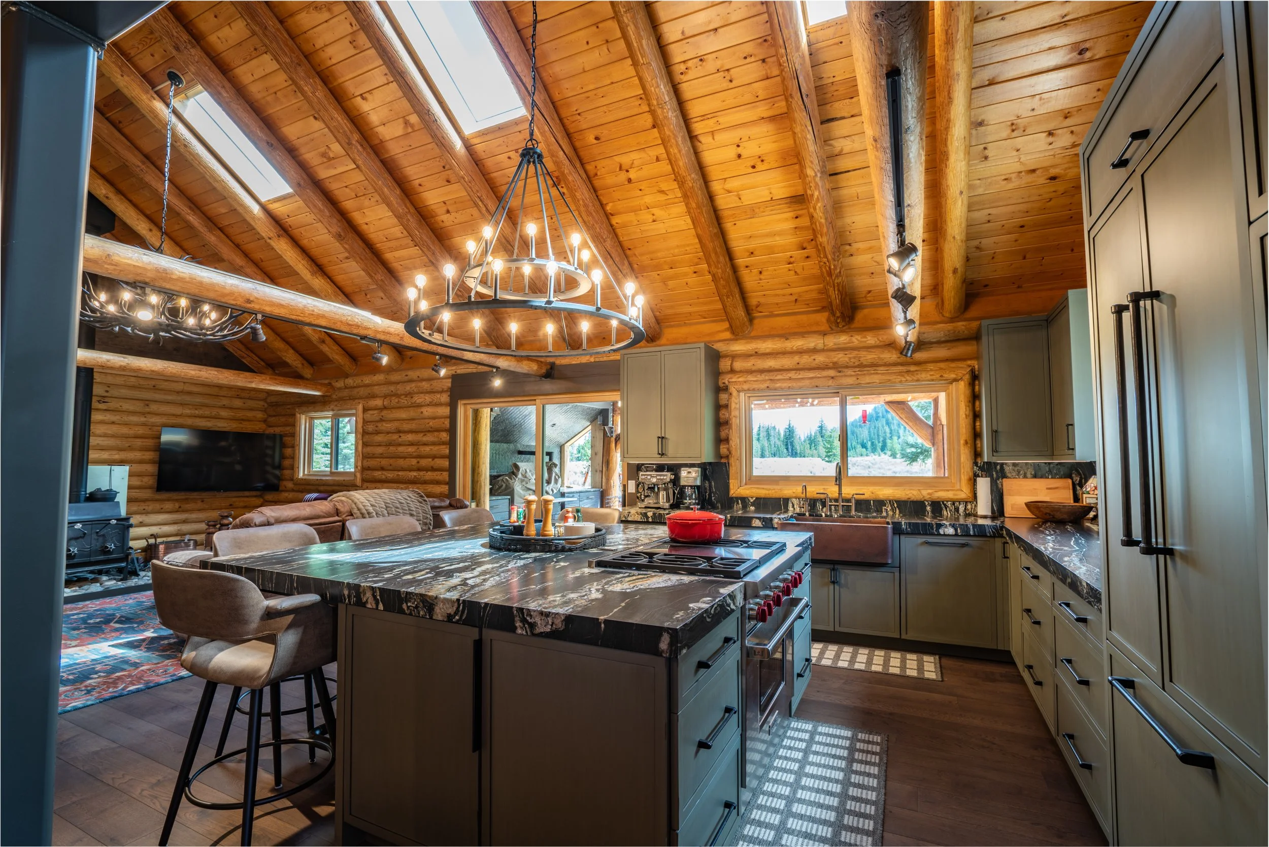 Interior view of a rustic kitchen with a large island, gray cabinets, and wood-paneled vaulted ceiling with skylights and a chandelier.
