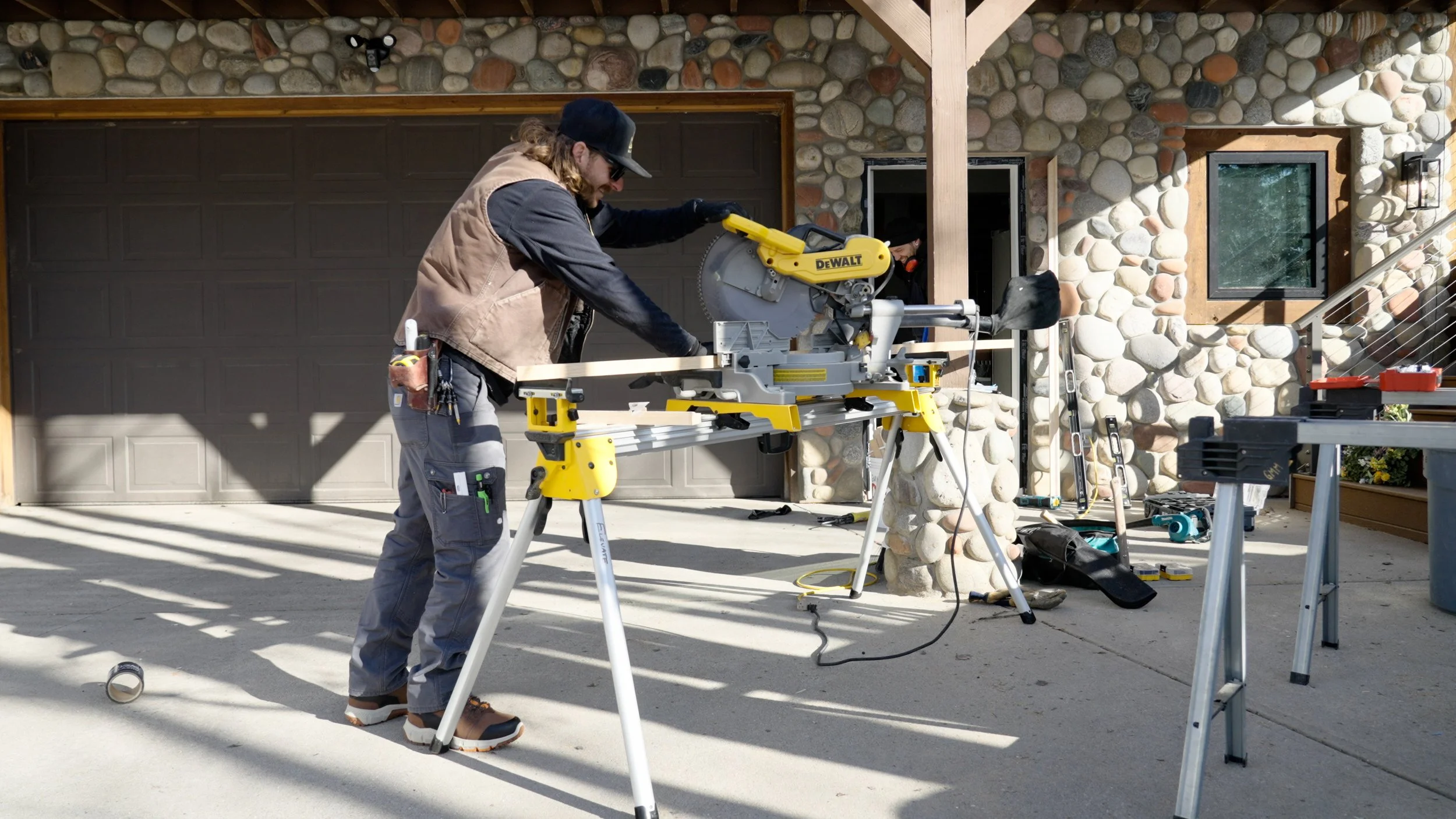 A man with long hair, beard, sunglasses, and a baseball cap operating a yellow DeWALT miter saw on a work table outside a house with a stone exterior wall. There are tools, a ladder, and equipment around him, and another person partially visible in the background.