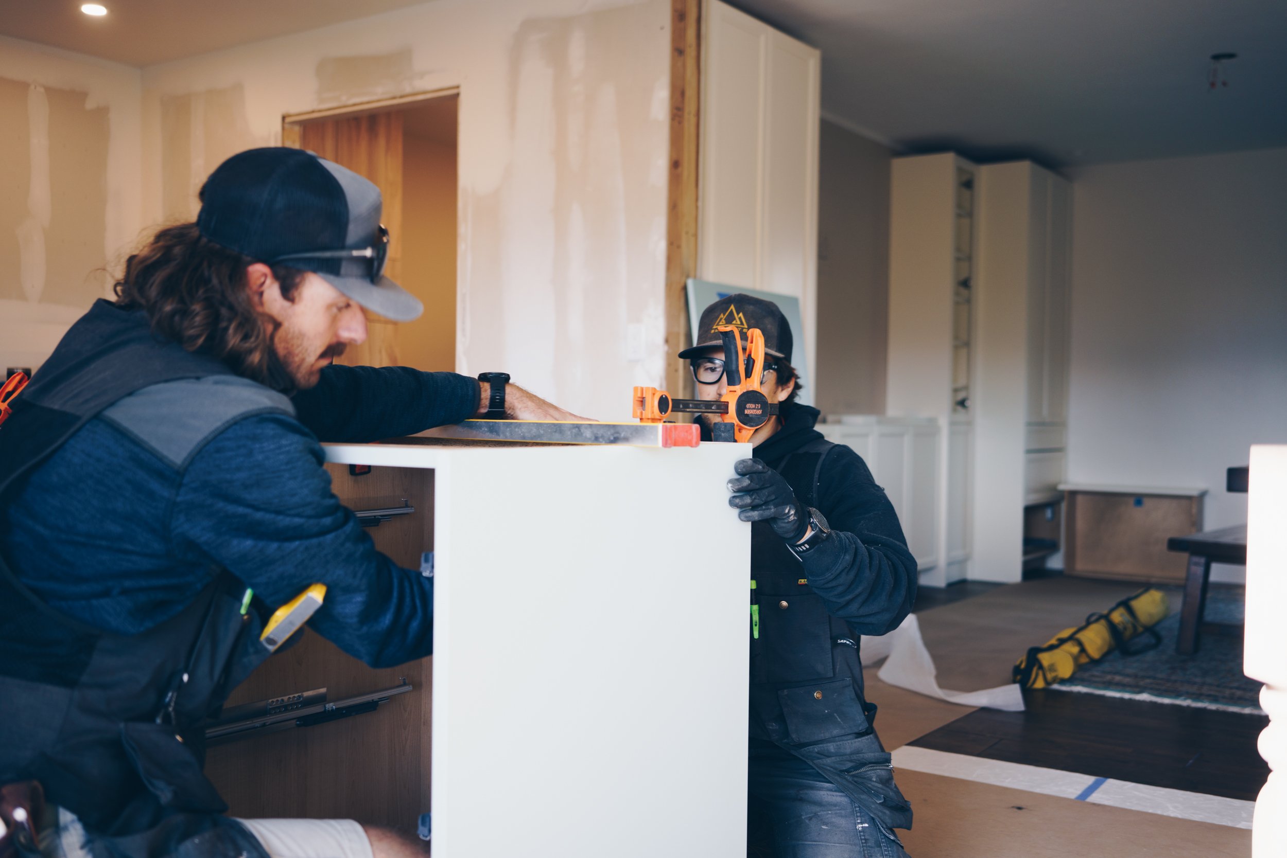 Two men are working together on a woodworking project inside a home. One man is wearing a blue hoodie and cap, and the other is wearing a black jacket, gloves, and a cap. They are using a tool to assemble or adjust a white cabinet or piece of furniture.