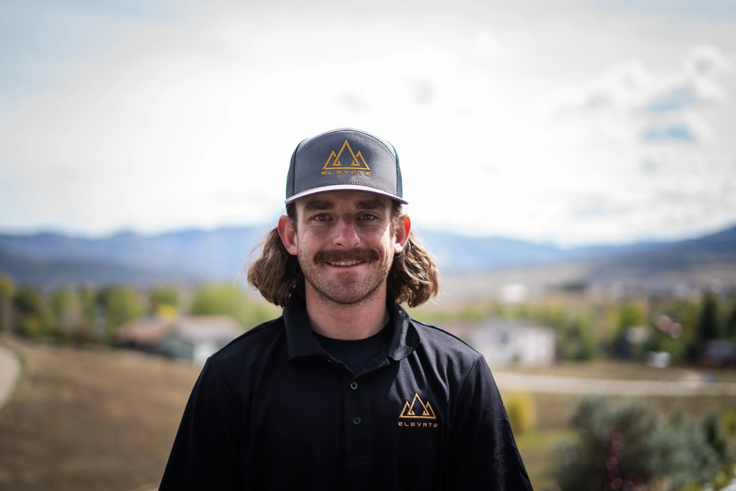 A man with long hair and a beard wearing a black shirt and a gray baseball cap with a gold mountain logo and the word "ELEVATE" stands outdoors against a background of mountains and cloudy sky.