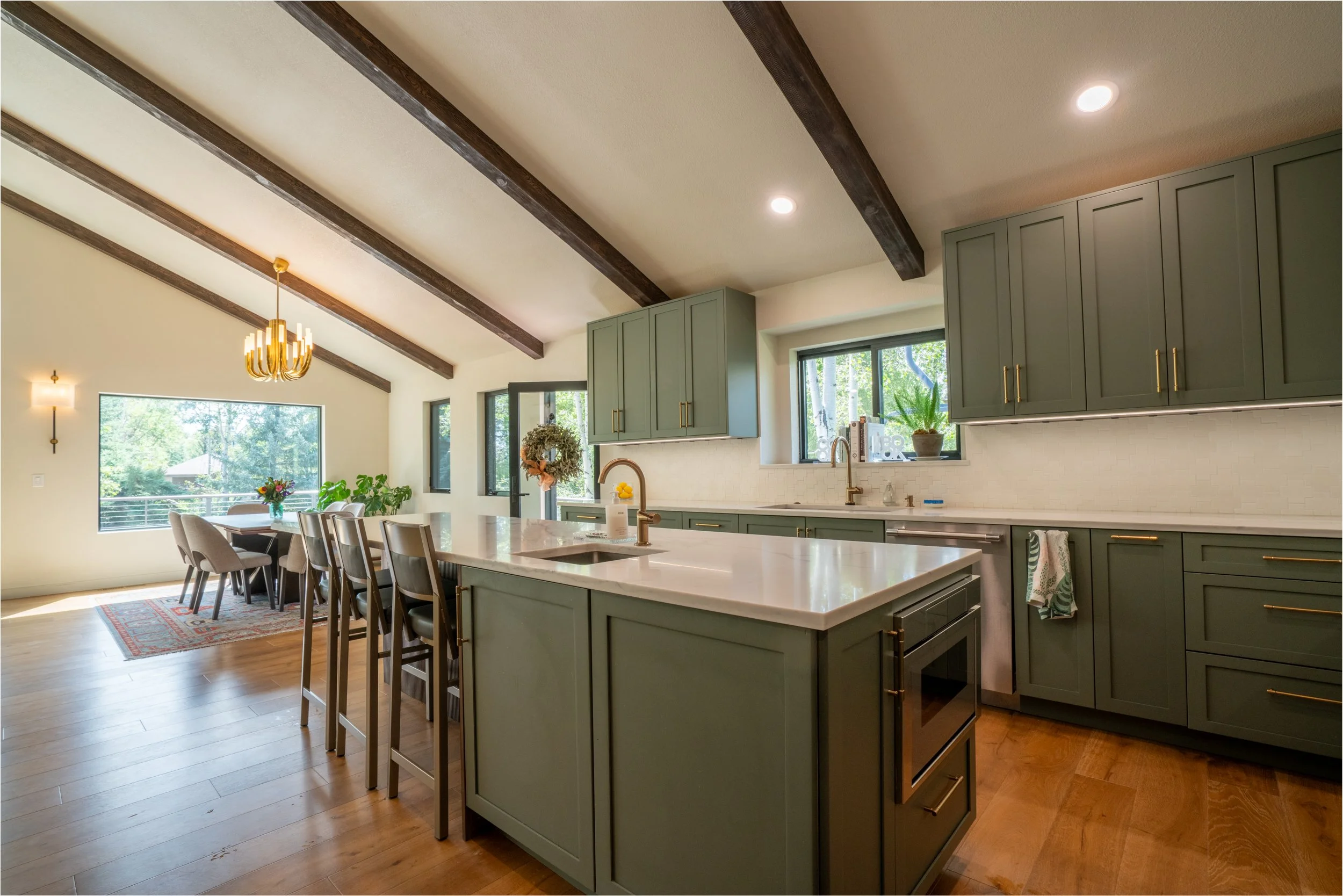 Modern kitchen with green cabinets, white countertop, pendant lighting, hardwood floors, large window showing outdoor greenery, and a dining area with a table and chairs.