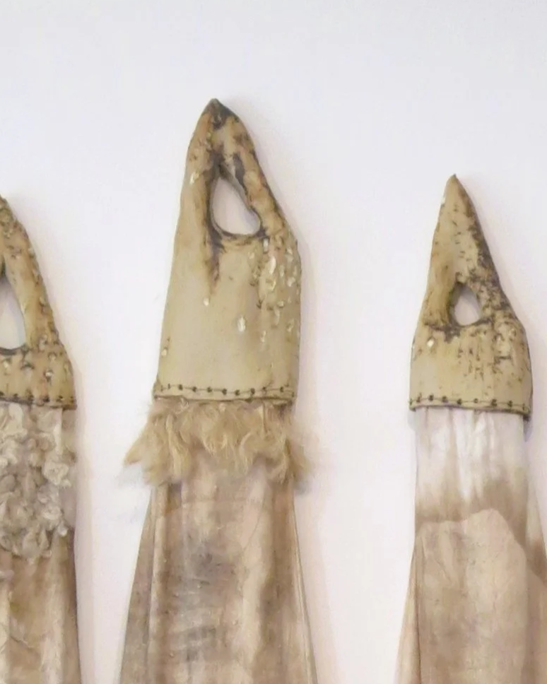 Trois masques ou sculptures en forme de visages humains avec des têtes en bois ou en tissu, exposés contre un mur blanc.