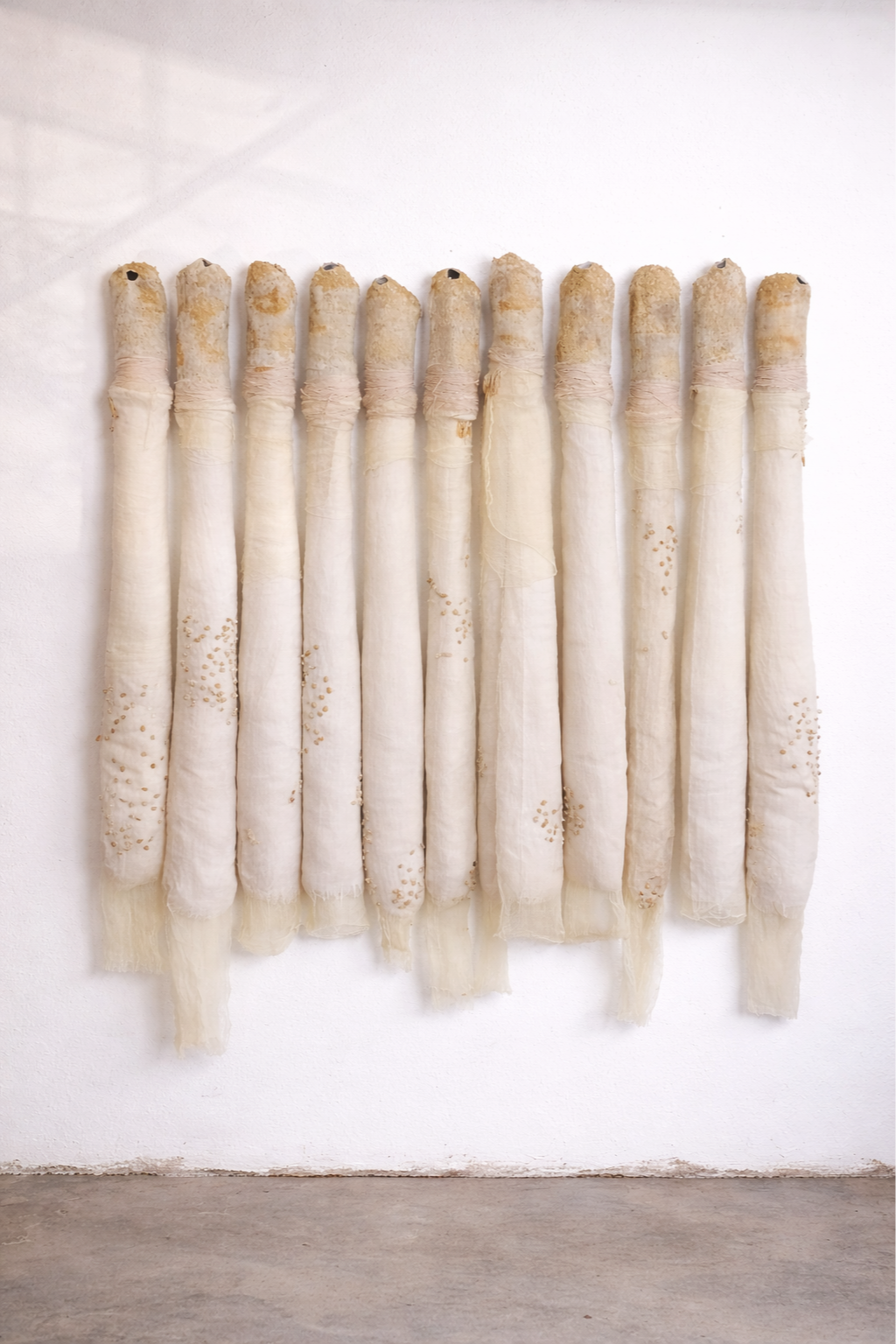 Une installation artistique composée de plusieurs longs tubes suspendus contre un mur blanc, ressemblant à des bandages ou des matériaux tissés.