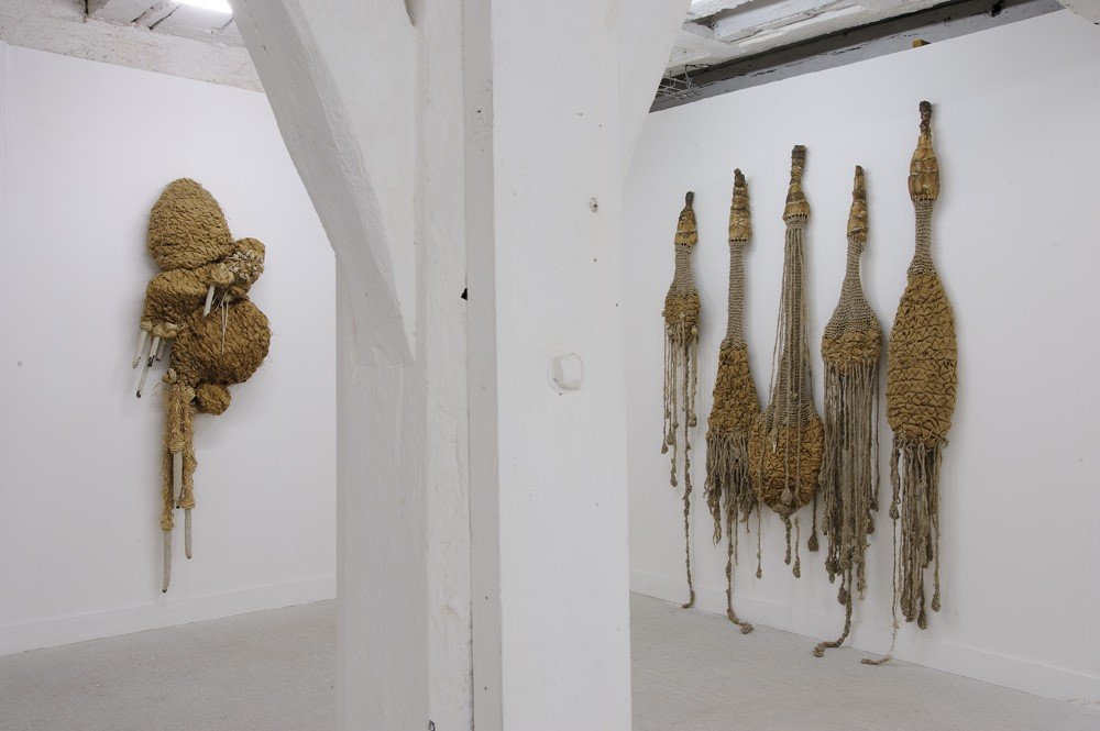 Installation d'œuvres en textile faites de matières naturelles comme la laine, suspendues sur un mur blanc, dans une galerie d'art