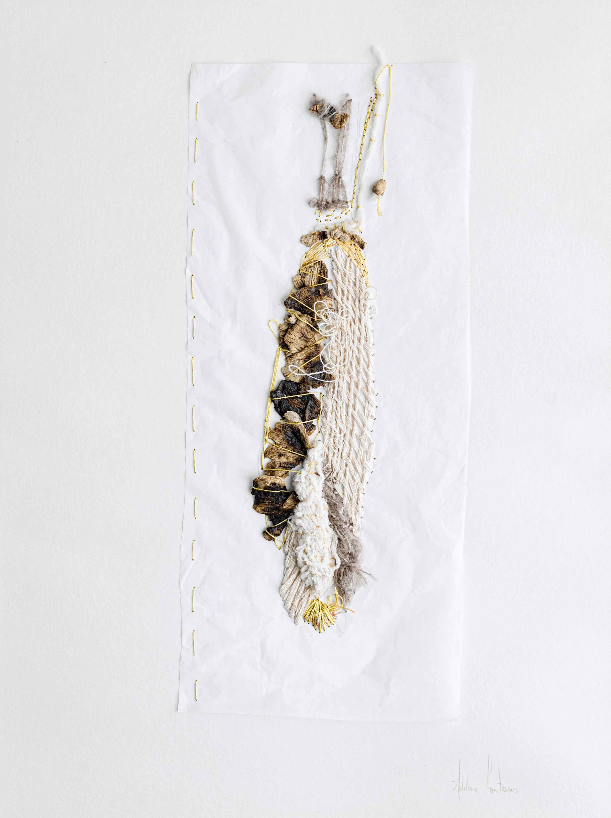 Une œuvre d'art faite avec des matériaux naturels comme des cailloux, des fils, de la corde, et des tissus, assemblés pour former une composition suspendue sur un fond blanc.