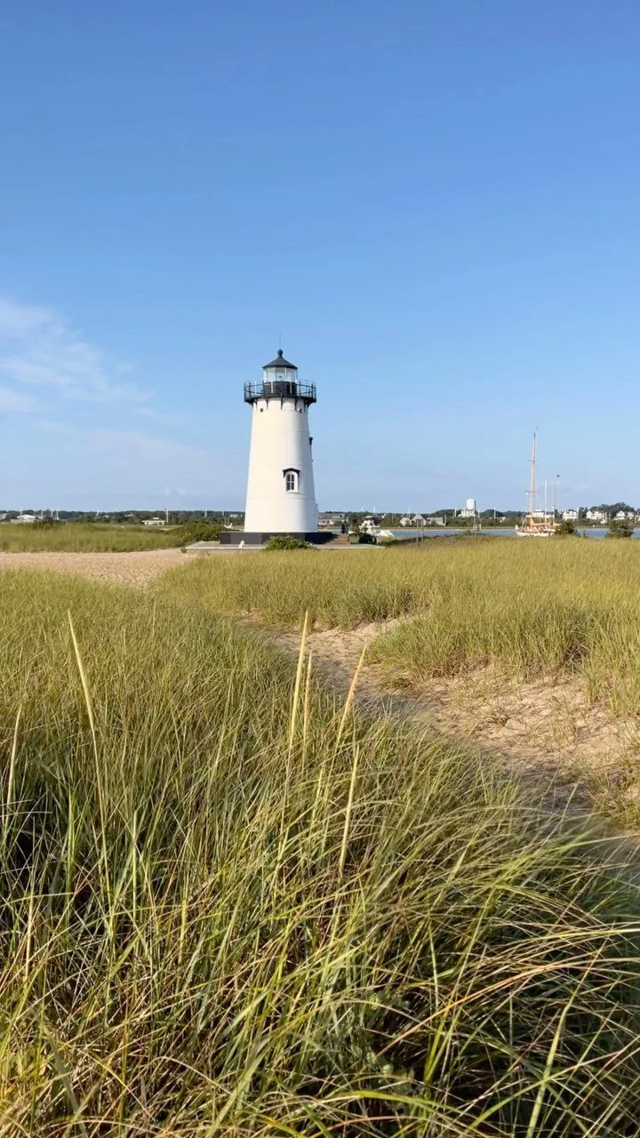 A lighthouse standing on a grassy landscape with a clear blue sky overhead and a sailboat in the distance.