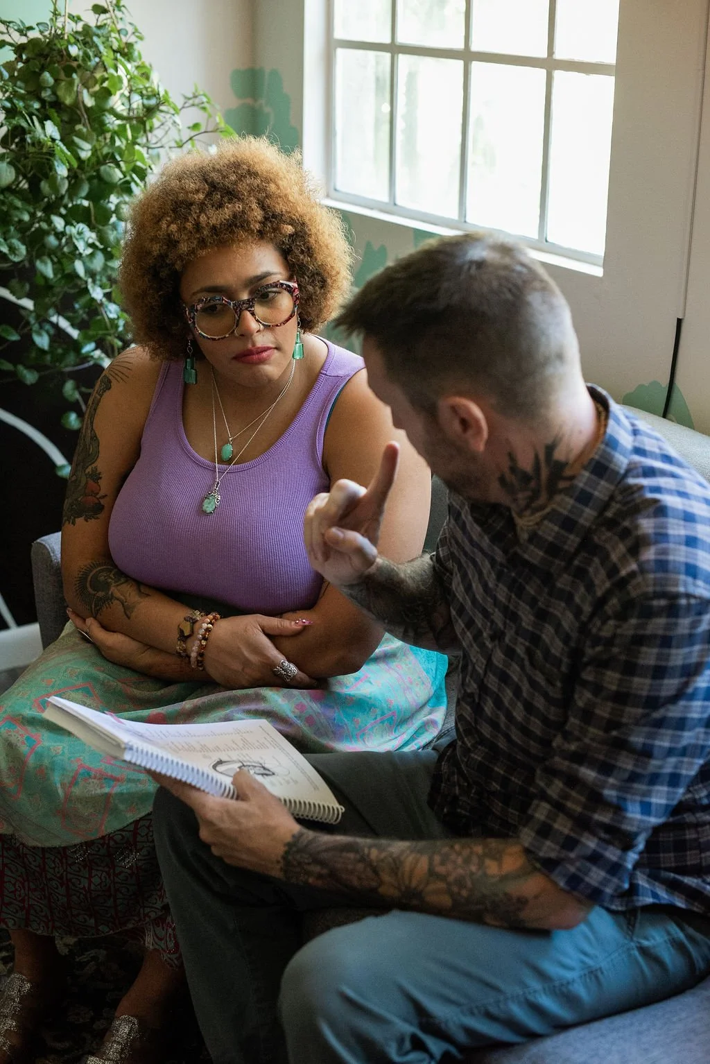 A woman with curly hair, glasses, and tattoos wearing a purple tank top and jewelry sits and listens, while a man with tattoos, short hair, and a plaid shirt shows her something in a notebook during a conversation in a bright room with a window and houseplant.