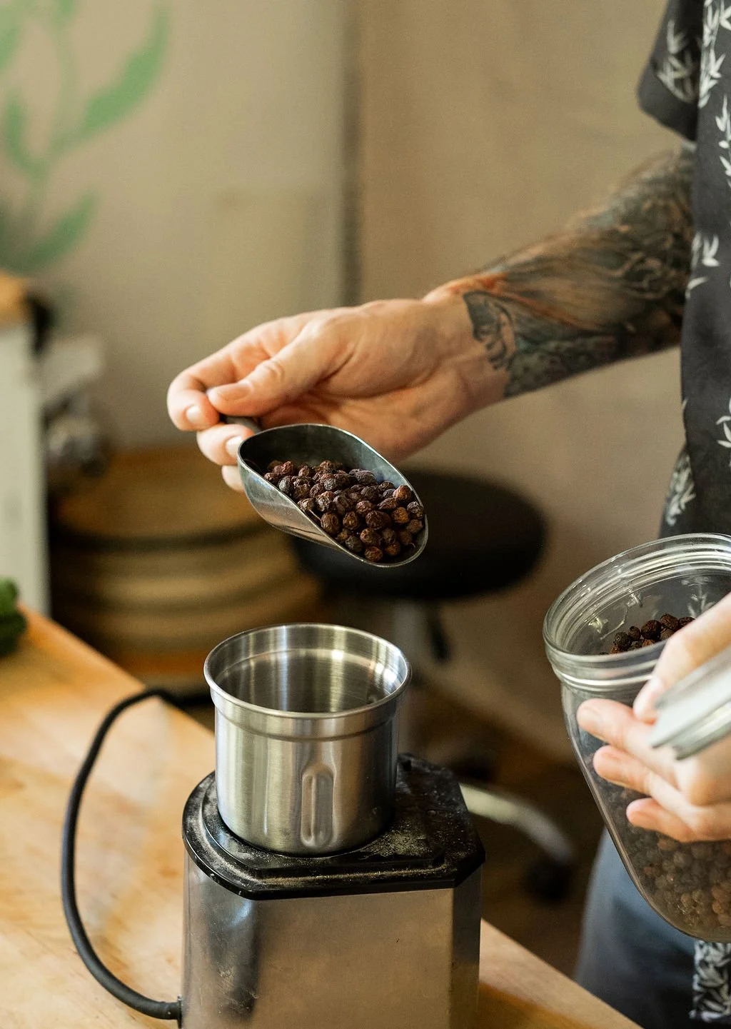 Person with tattooed arm pouring black peppercorns into a metal container near an electric grinder on a wooden table.
