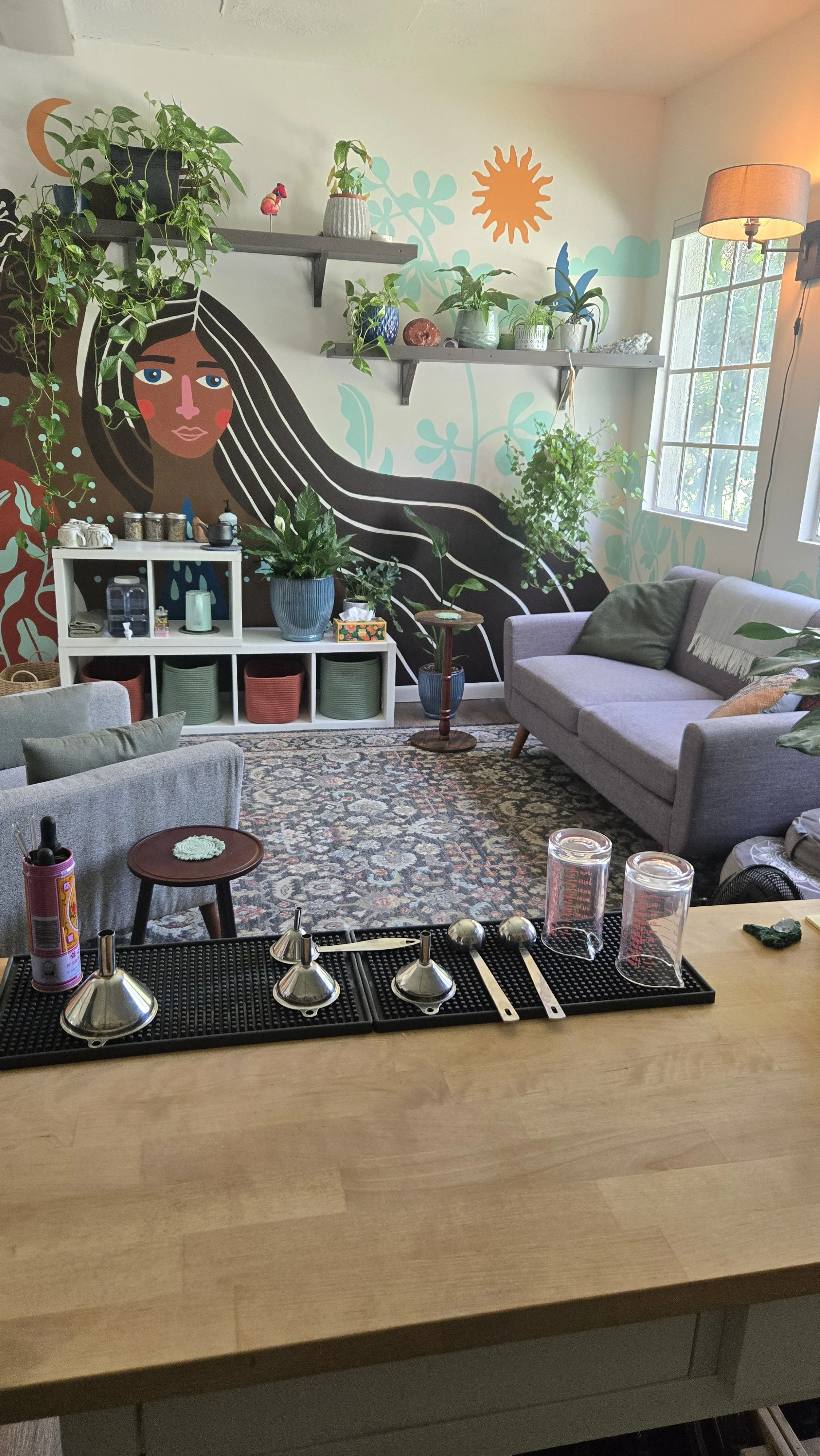 Coffee bar countertop with silver bells, glass cups, and utensils in a cozy living room with a colorful mural and potted plants.