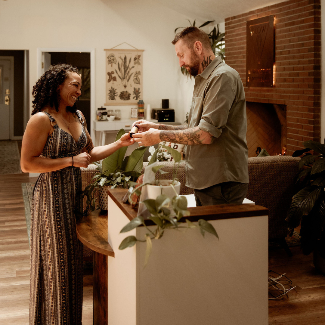 A man is giving a woman a small object while she smiles and reacts happily, standing in a cozy, warmly lit living room with plants and artwork.