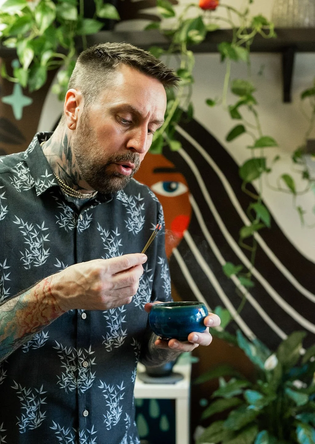 A man with tattoos on his arms, styled hair, and tattoos on his neck, is holding a small brush and a blue ceramic bowl. He is wearing a black shirt with white leaf patterns and appears to be focused on a task. The background features a colorful mural with an abstract face and hanging green plants.