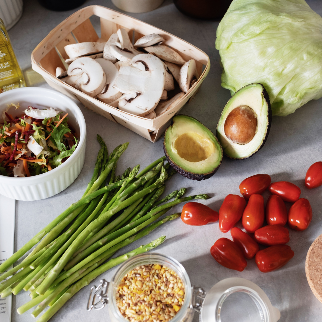 Fresh vegetables including sliced mushrooms in a basket, sliced avocado halves, cherry tomatoes, a bunch of asparagus, shredded carrots and cabbage salad in a bowl, a head of lettuce, and a jar of chopped nuts.