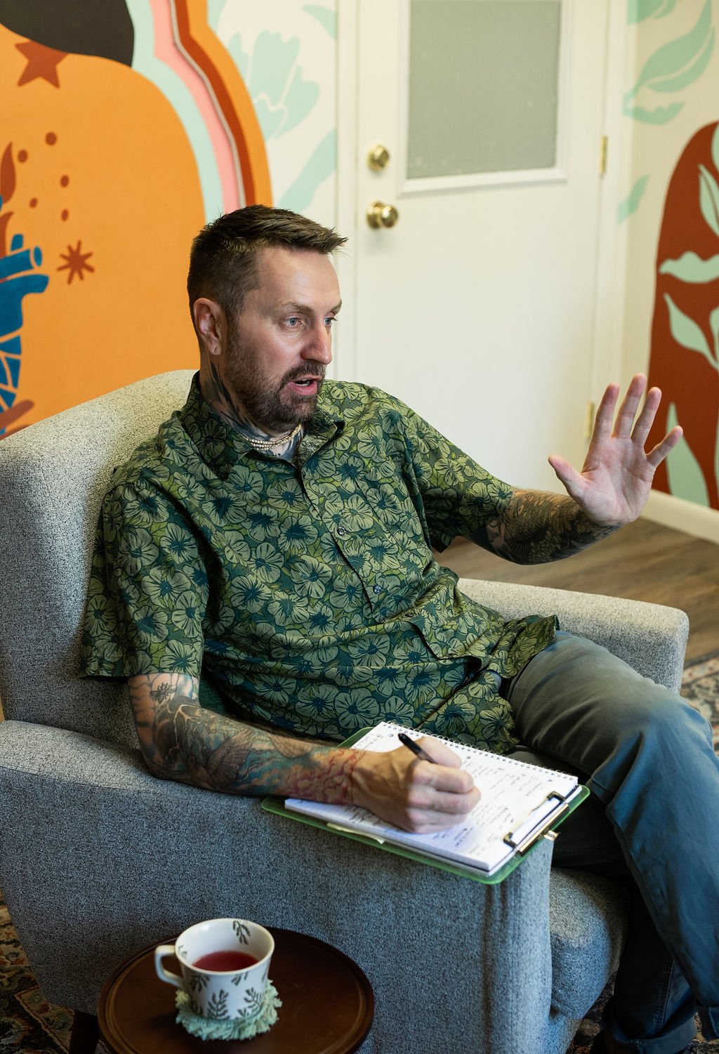 A man with tattoos, wearing a patterned shirt, seated on a gray armchair, writing on a clipboard, with a cup of tea on a small table nearby, in a room with colorful artwork on the walls.