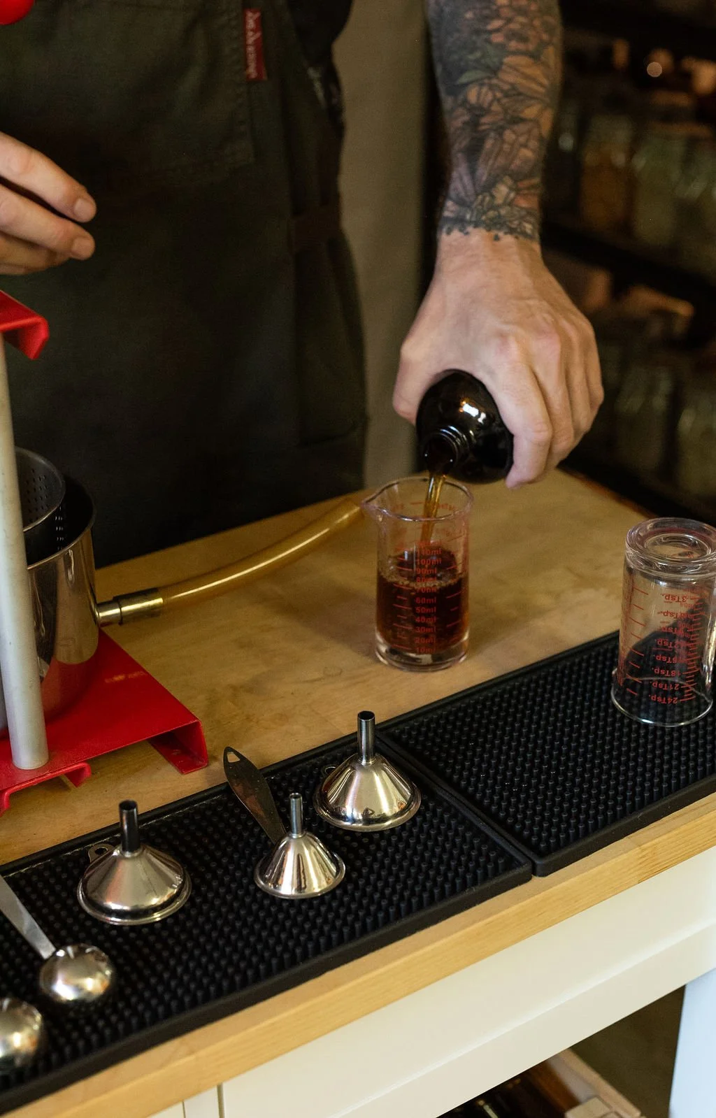 Person pouring dark liquid from a bottle into a measuring glass on a bar counter, with various bar tools and measurement cups nearby.