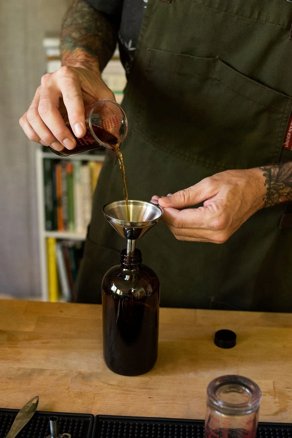 A person with tattooed arms wearing a dark apron is pouring a dark liquid from a measuring cup into a metal funnel placed inside a bottle on a wooden surface.