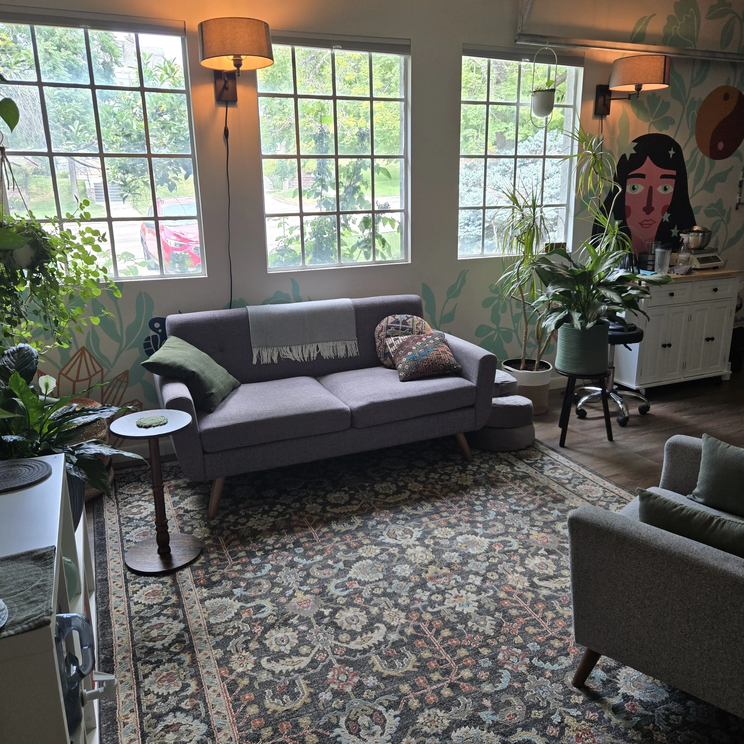 A living room with large windows, a patterned rug, a gray sofa with colorful pillows, a small round side table, various potted plants, and a decorative mural on the wall with an illustration of a woman's face and green foliage.