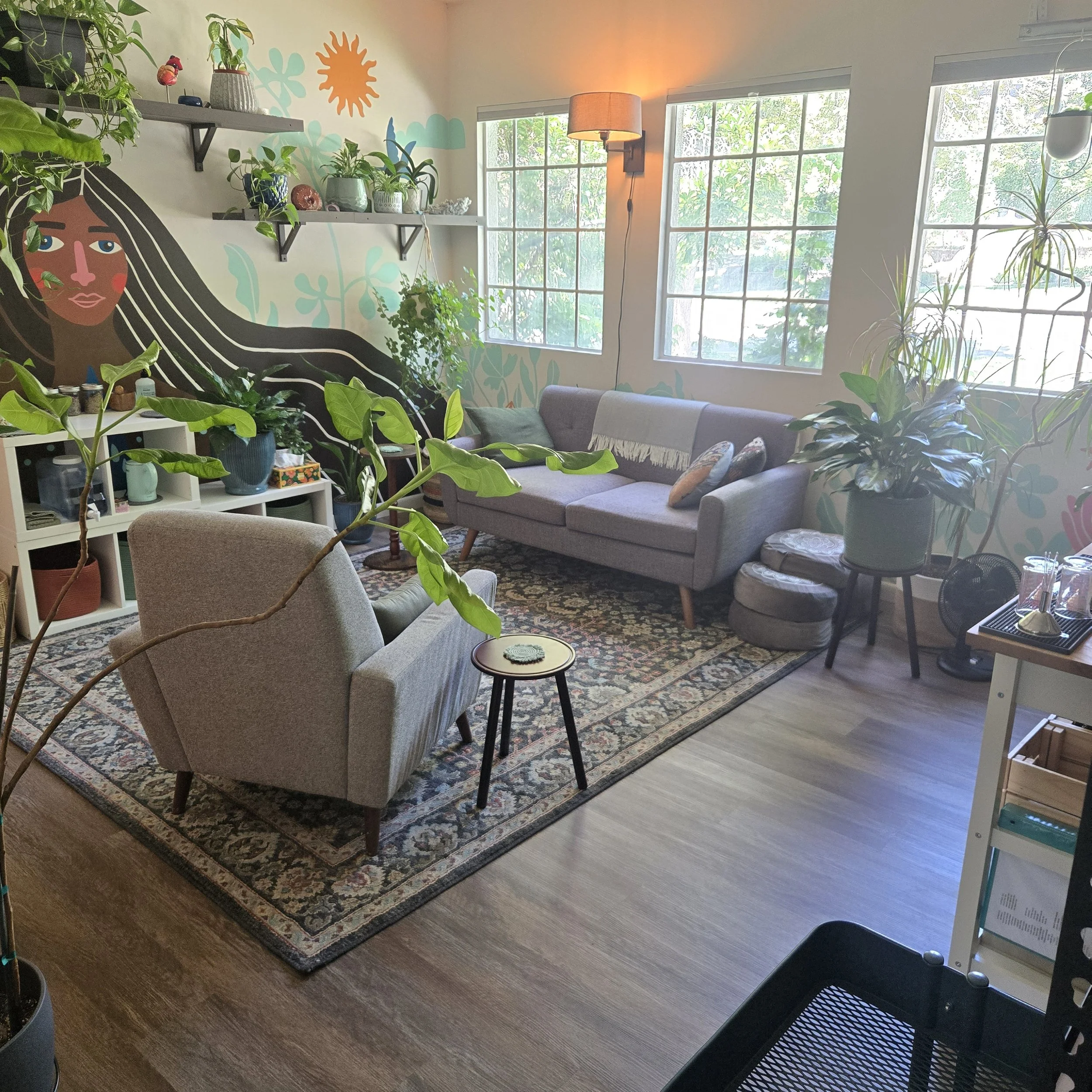 A cozy living room with large windows letting in natural light, decorated with plants, a mural of a woman's face and hair on the wall, a standing lamp, a gray sofa with pillows, a patterned rug, and various shelves and furniture.