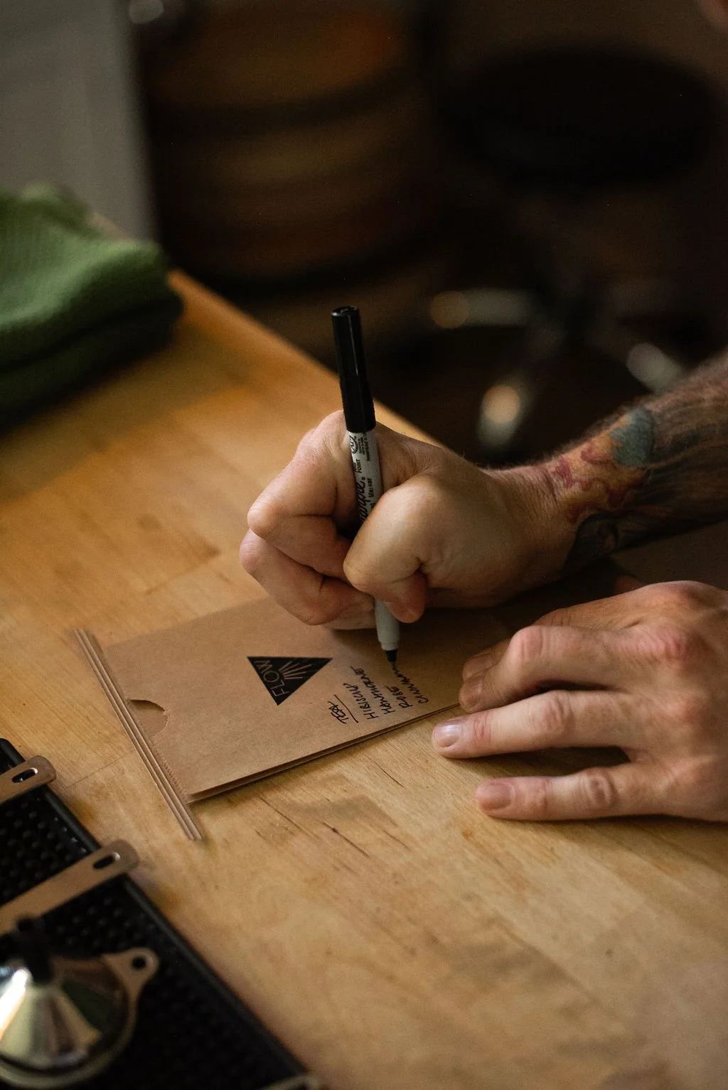A person with tattoos on their arm writing on a small brown paper bag with a black marker pen on a wooden table.