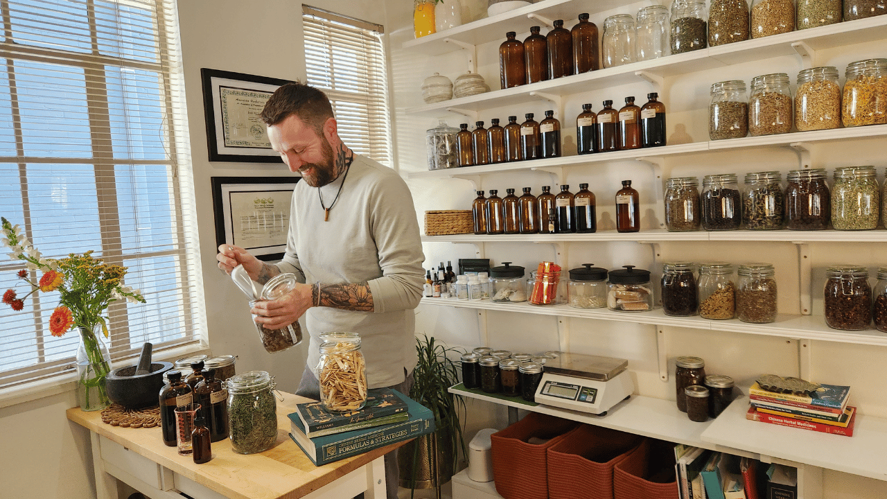 A man with tattoos on his arms, beard, and short hair in a light-colored shirt, standing at a wooden table in a bright room. He is holding a jar filled with herbs or dried plants. The room has shelves filled with various jars and bottles containing herbs, dried plants, and powders, as well as scales and books. A bouquet of orange and white flowers is on the table near a mortar and pestle.