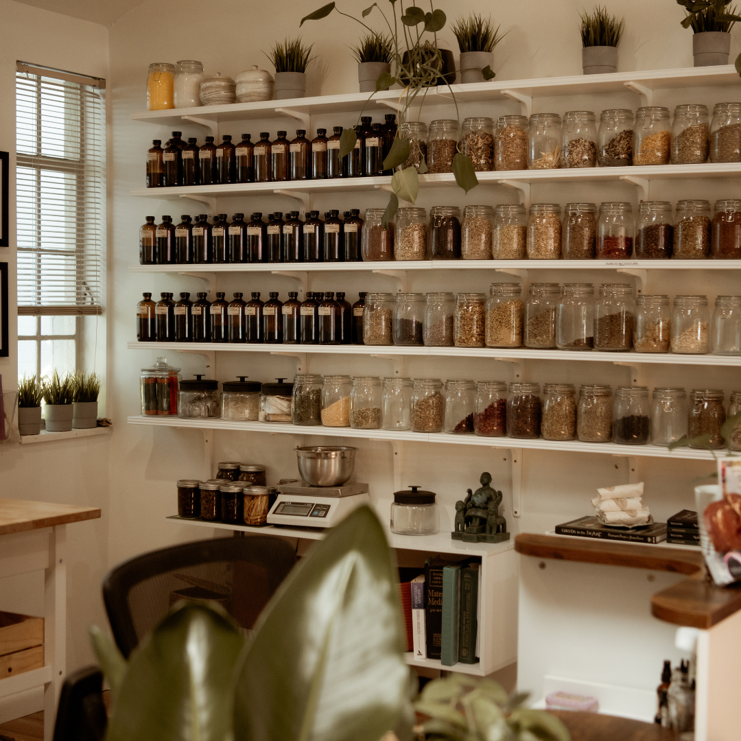 A home spice cabinet with shelves filled with jars of dried herbs and spices, potted plants on top, and some books and kitchen items nearby.