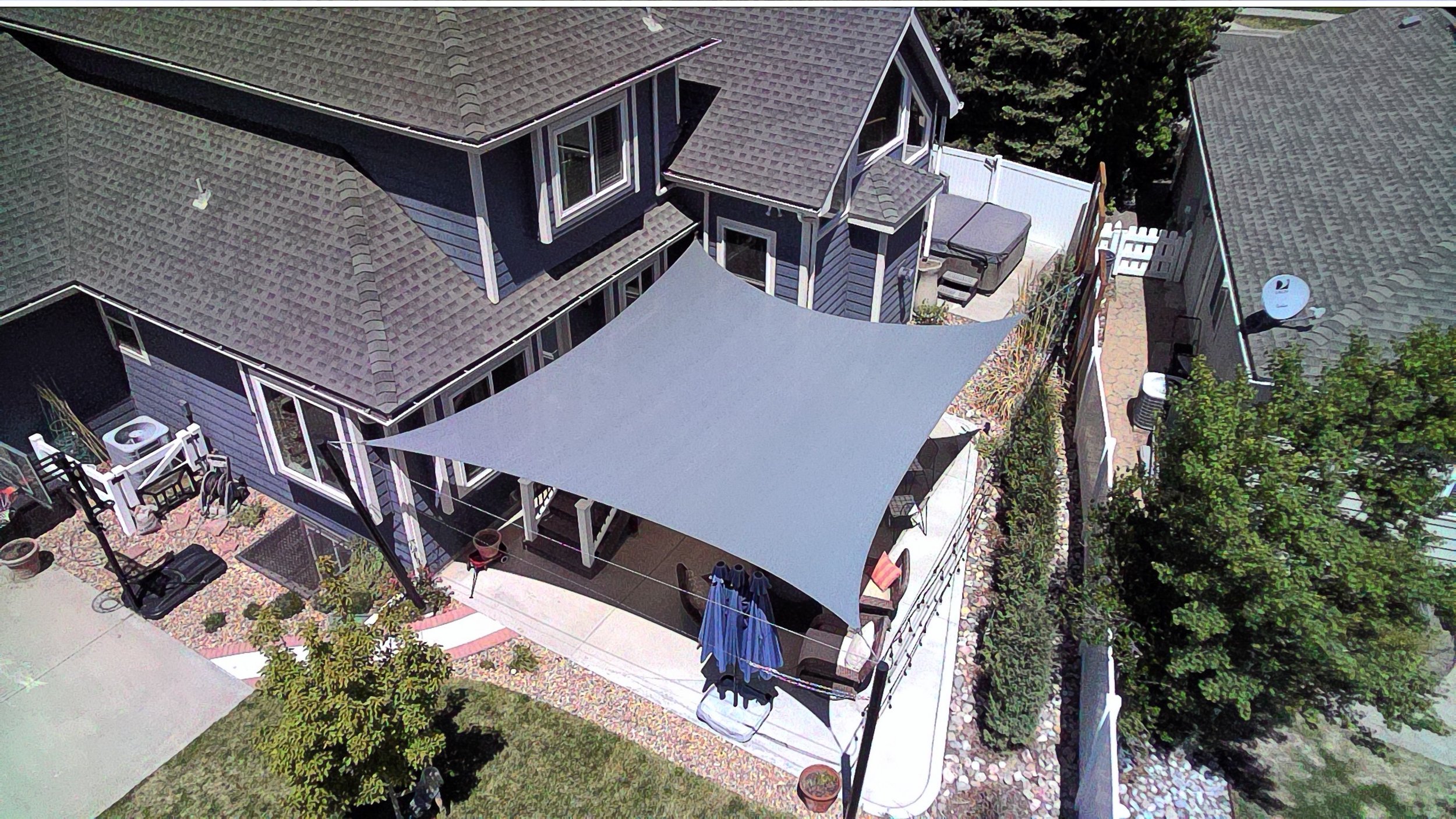 Aerial view of a house backyard with a large gray shade sail, patio furniture, potted plants, landscaping, and neighboring houses.