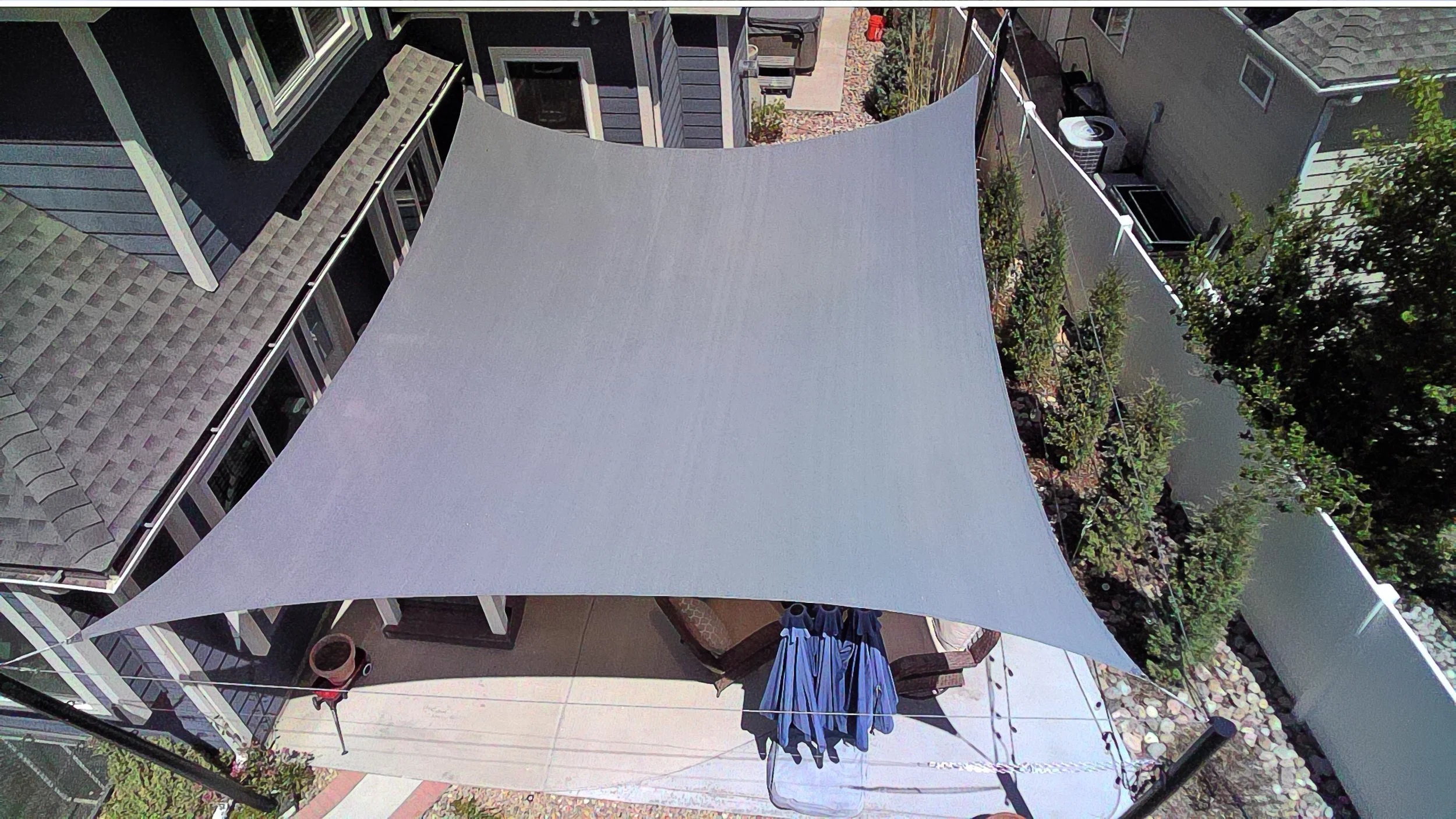 Backyard patio area with a large shade sail, outdoor furniture, potted plants, and neighboring houses with various outdoor items.