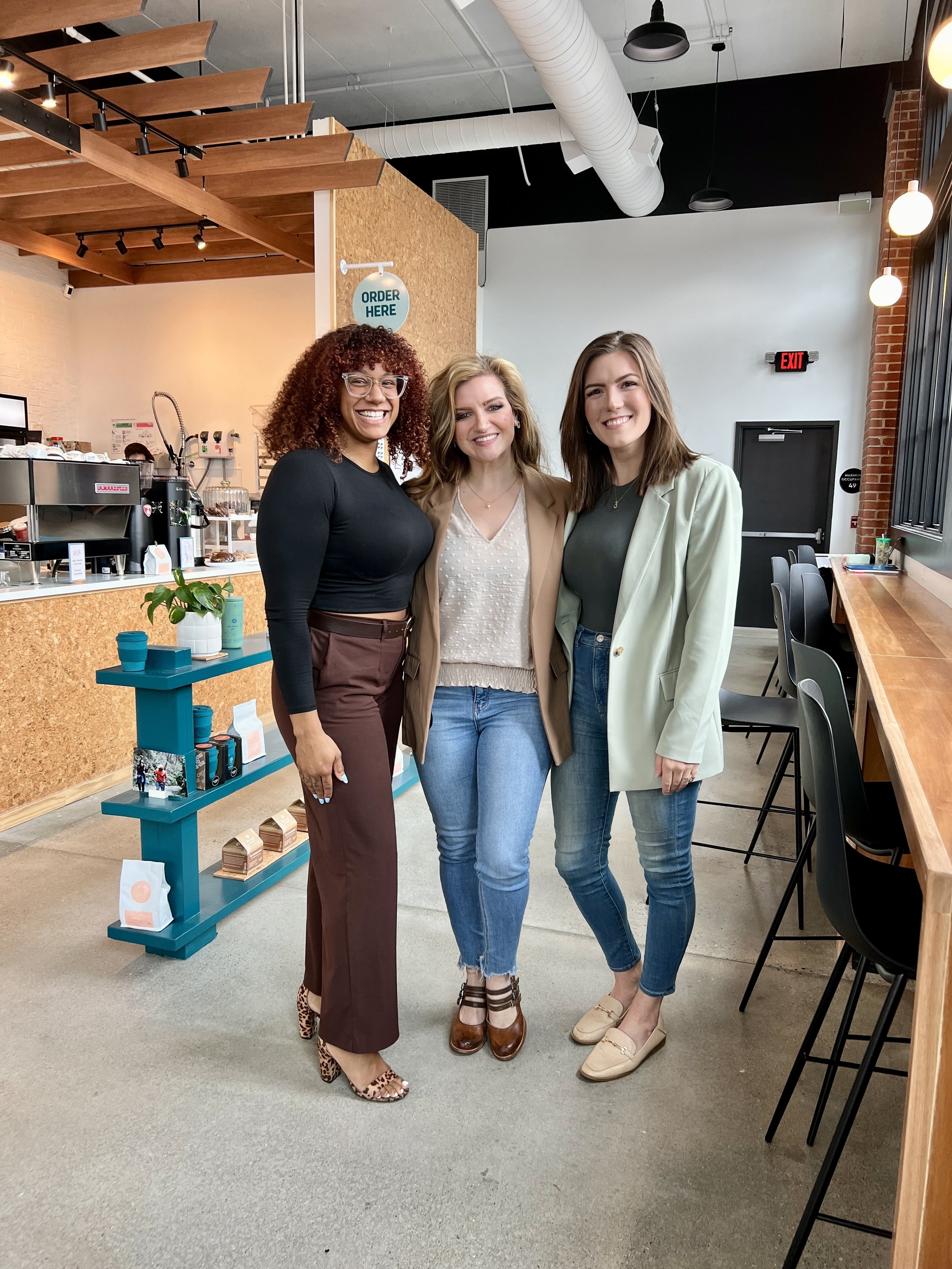 Three women smiling and standing in a modern cafe, next to a wooden counter and a coffee machine, with shelves displaying products in the background.
