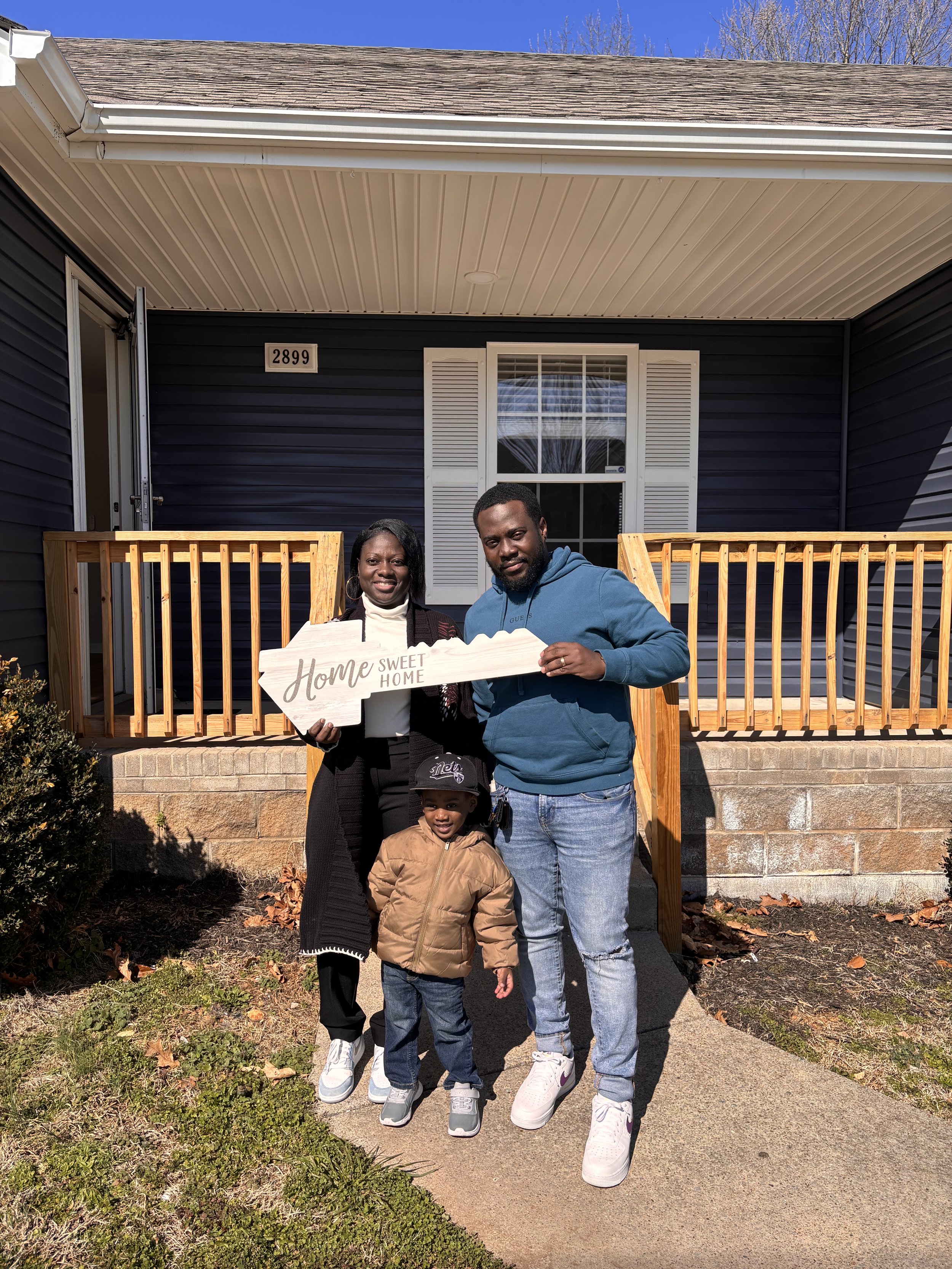 A family of three stands in front of a house holding a "Home Sweet Home" sign. The house has a blue exterior and a white porch railing. The family includes two adults and a child, all smiling. The house address is visible above the entrance.