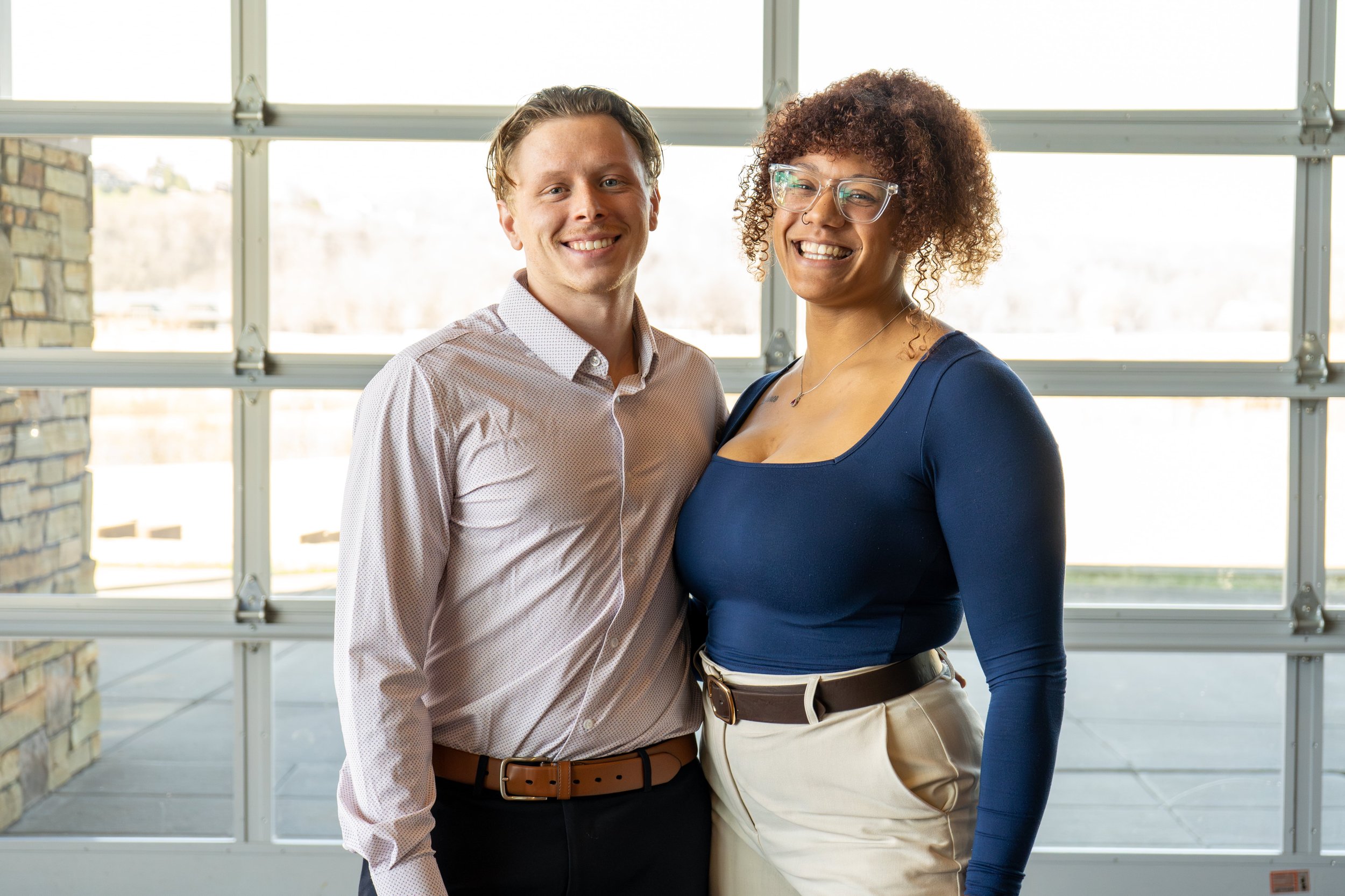 Two people standing and smiling indoors near a window, one wearing a light-colored shirt and the other in a dark top.