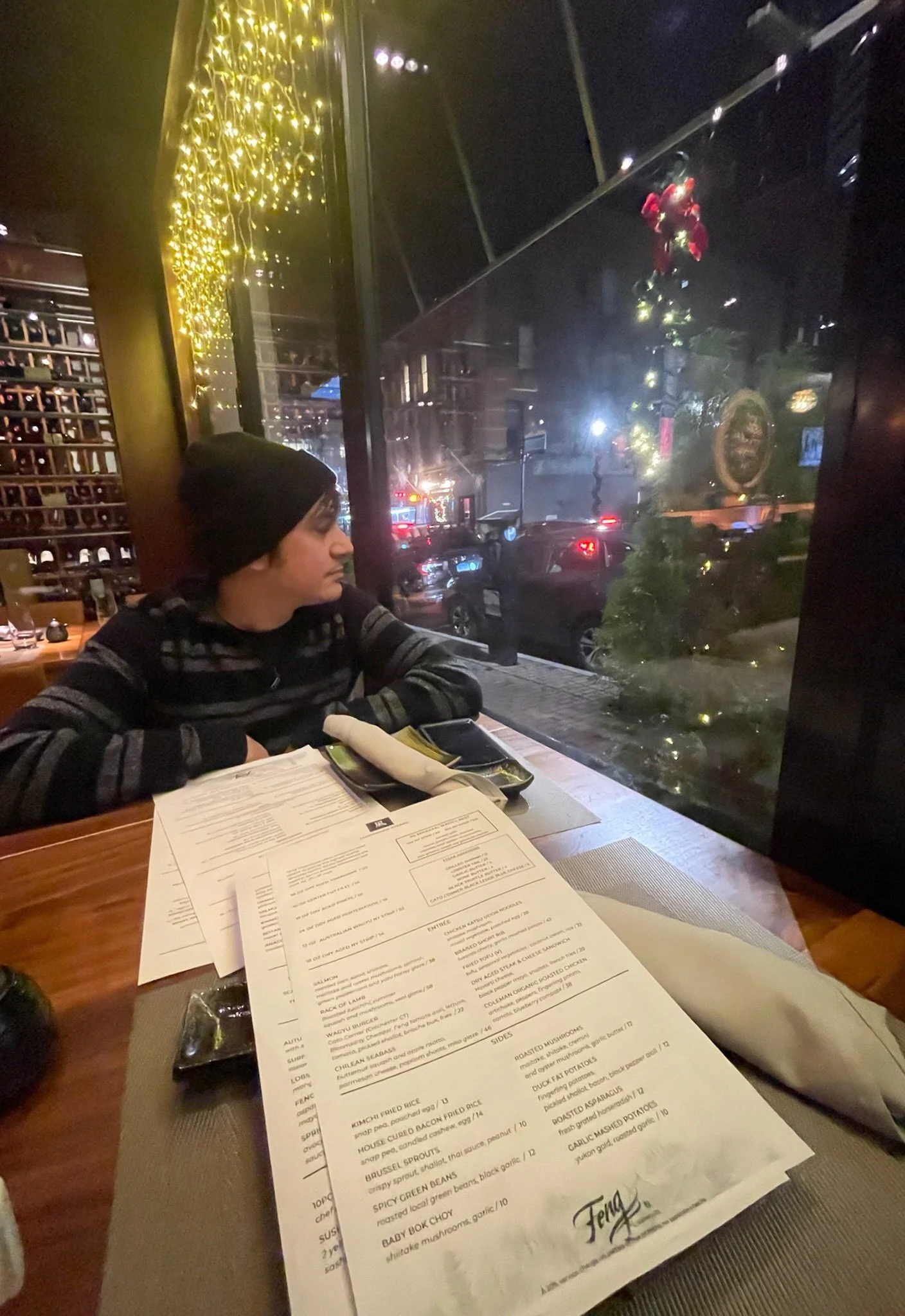 Person wearing a beanie sitting at a restaurant table looking out the window at night, with a menu and a napkin on the table; city lights and a Christmas tree visible outside.