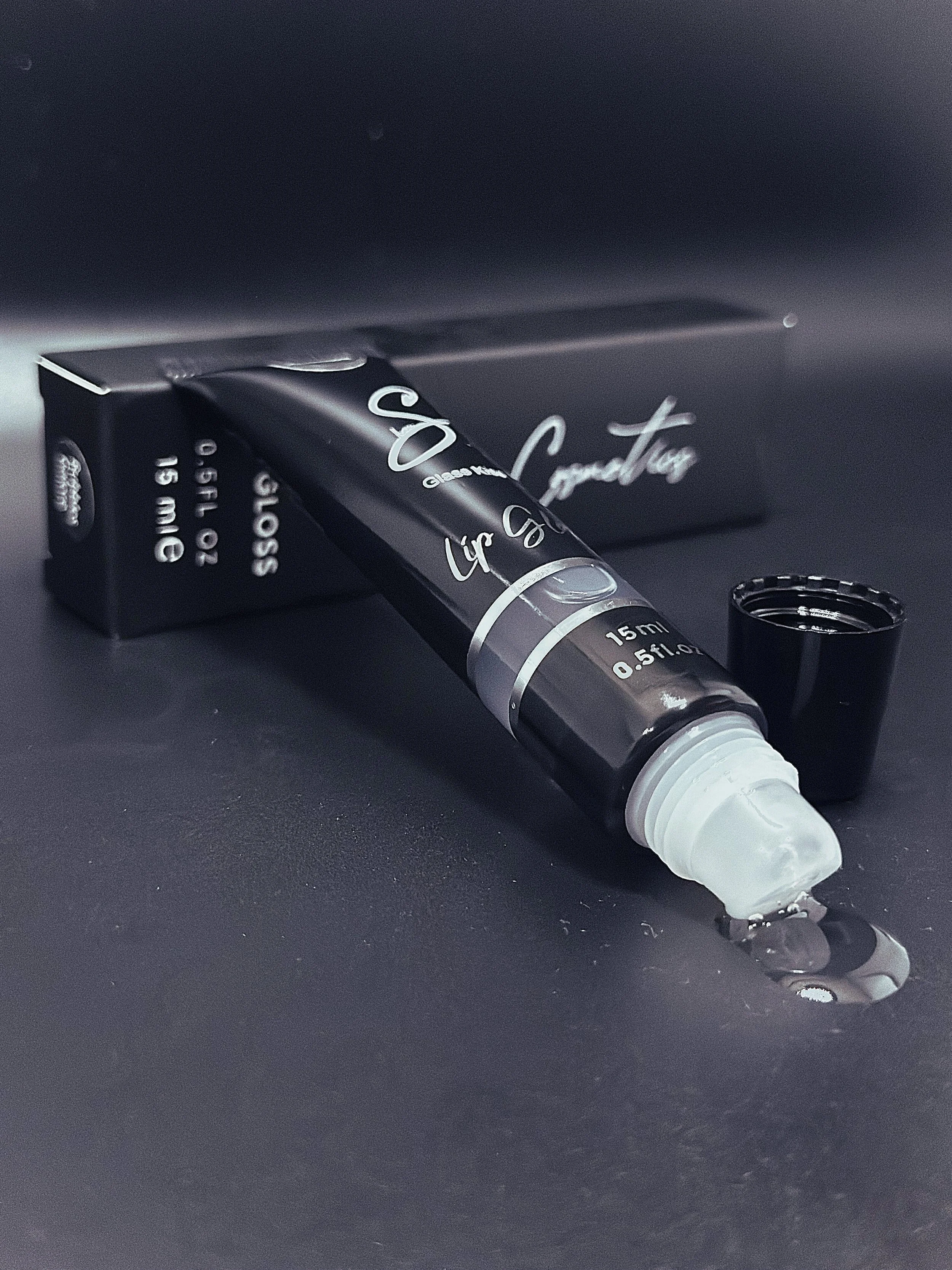 Lip gloss tube with clear lip gloss and top placed beside it on a black surface.