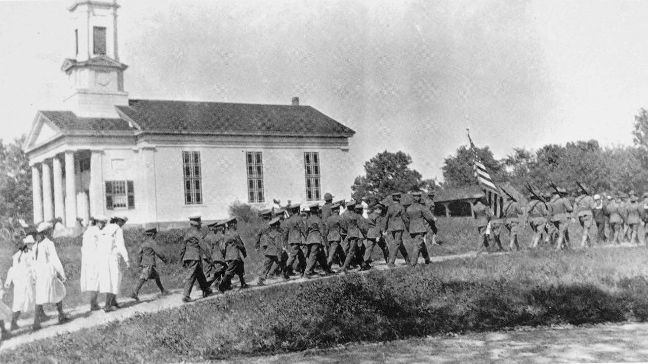 A historical black and white photograph of a group of African American soldiers marching with a flag near a white building with a clock tower.