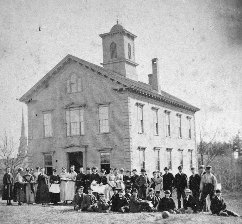 Historical black-and-white photo of a large two-story brick schoolhouse with a cupola, surrounded by a group of boys and girls standing and sitting in front.