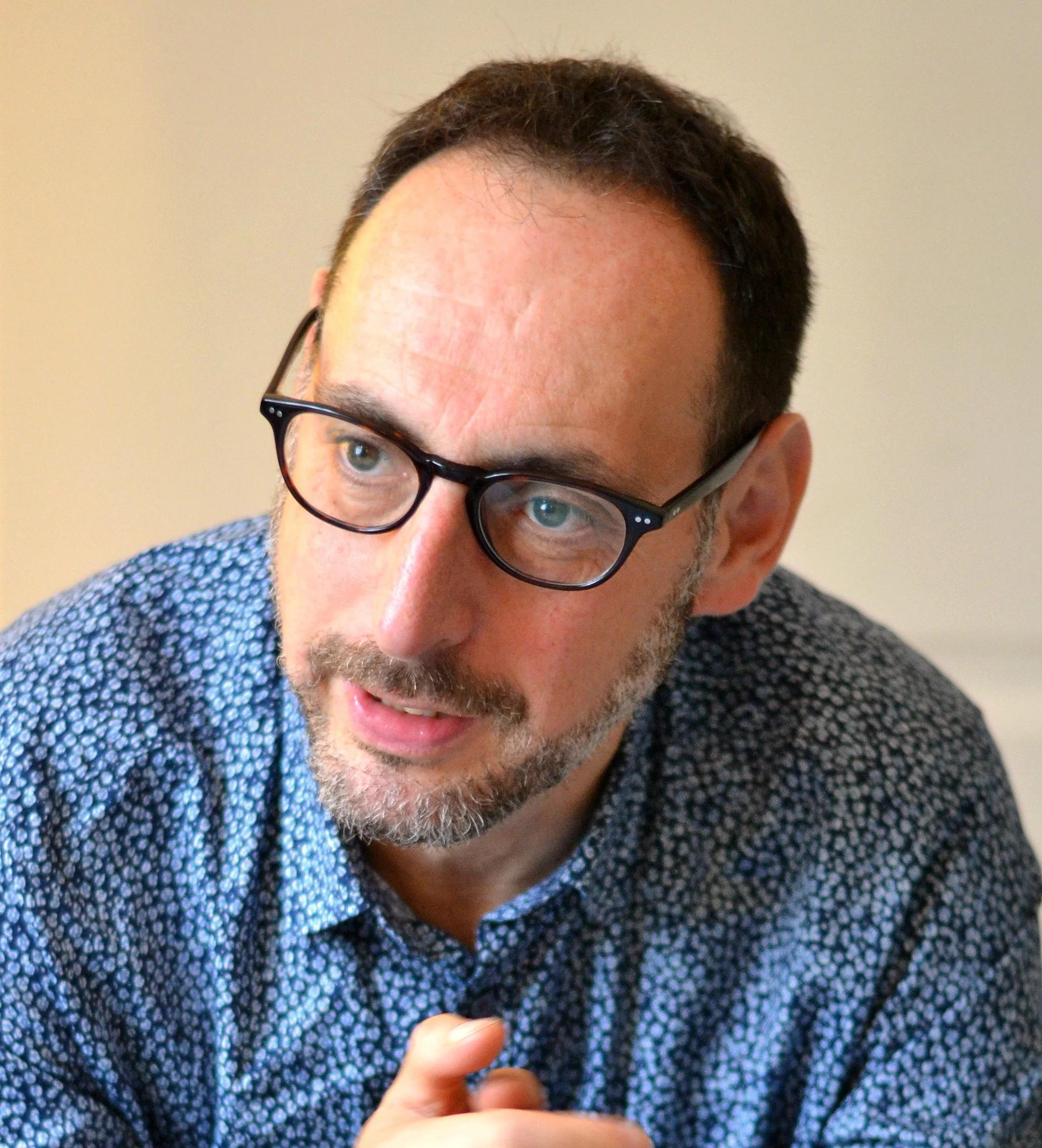 A man with short dark hair, glasses, and a beard, wearing a blue patterned shirt, is looking to his right during a conversation indoors.
