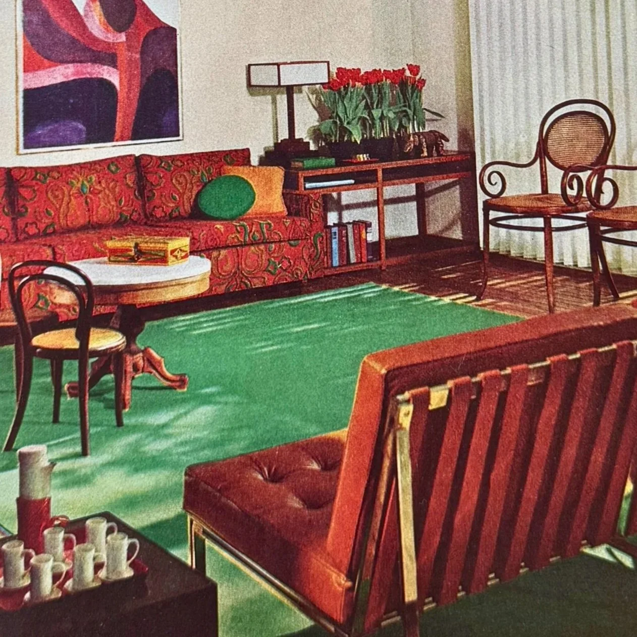 Vintage living room with red and green decor, featuring a patterned sofa, abstract wall art, red flowers on a side table, and a green rug.