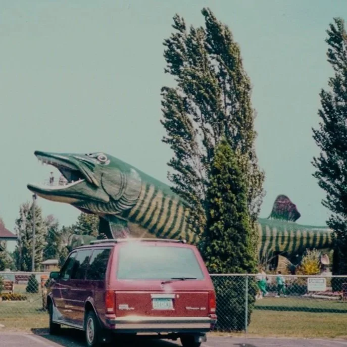 A large fish statue behind a red minivan in a park-like setting with trees and fences, captioned "MAKE NEW MEMS."