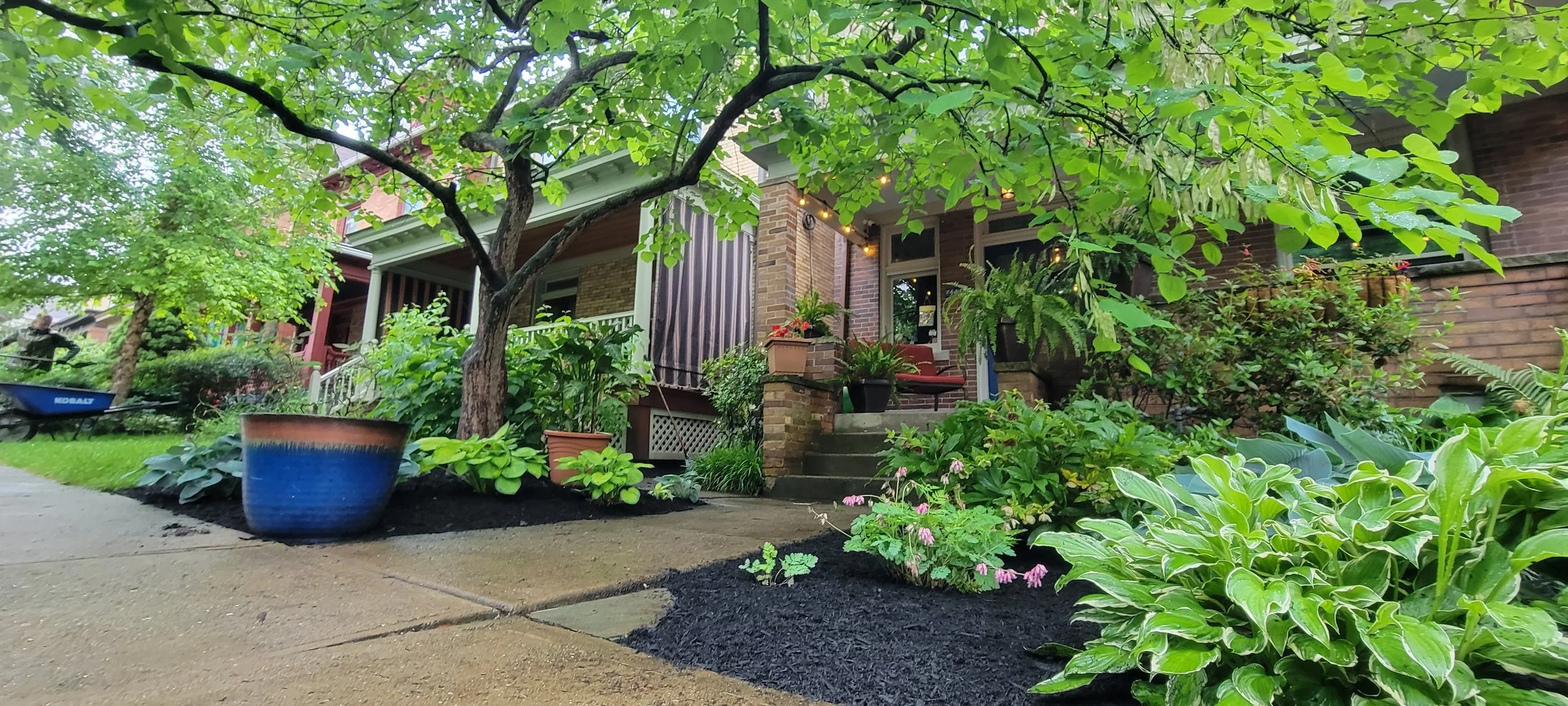 Front yard garden with lush greenery, potted plants, and a small pathway leading to a porch with a seating area.